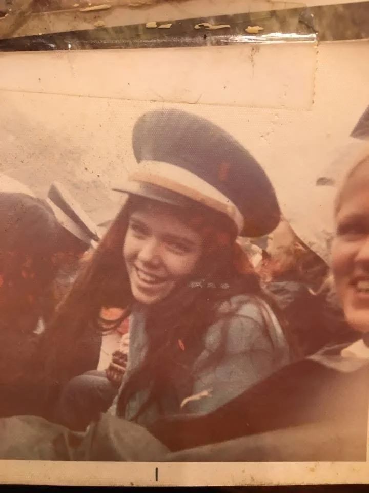A young woman with long brown hair smiling, wearing a black and white cap, in a group setting.