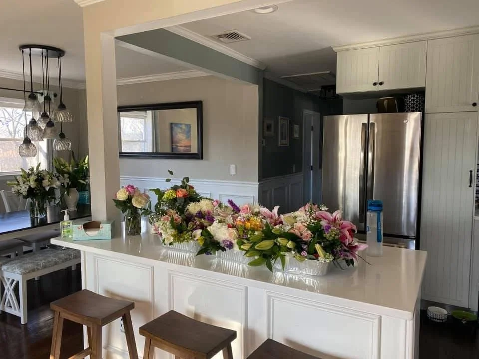 Kitchen island with arrangements of flowers in various vases, stainless steel refrigerator, white cabinets, and a glimpse of the dining area with hanging light fixtures.