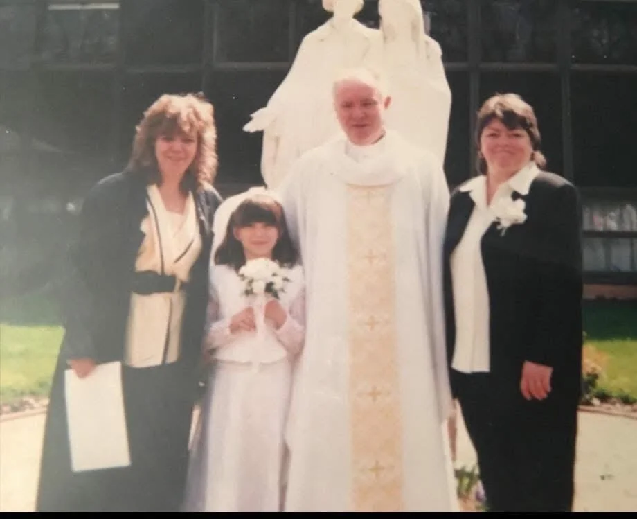A religious wedding scene featuring a priest and four women, one young girl, all in formal attire, standing outdoors in front of a building with large windows.