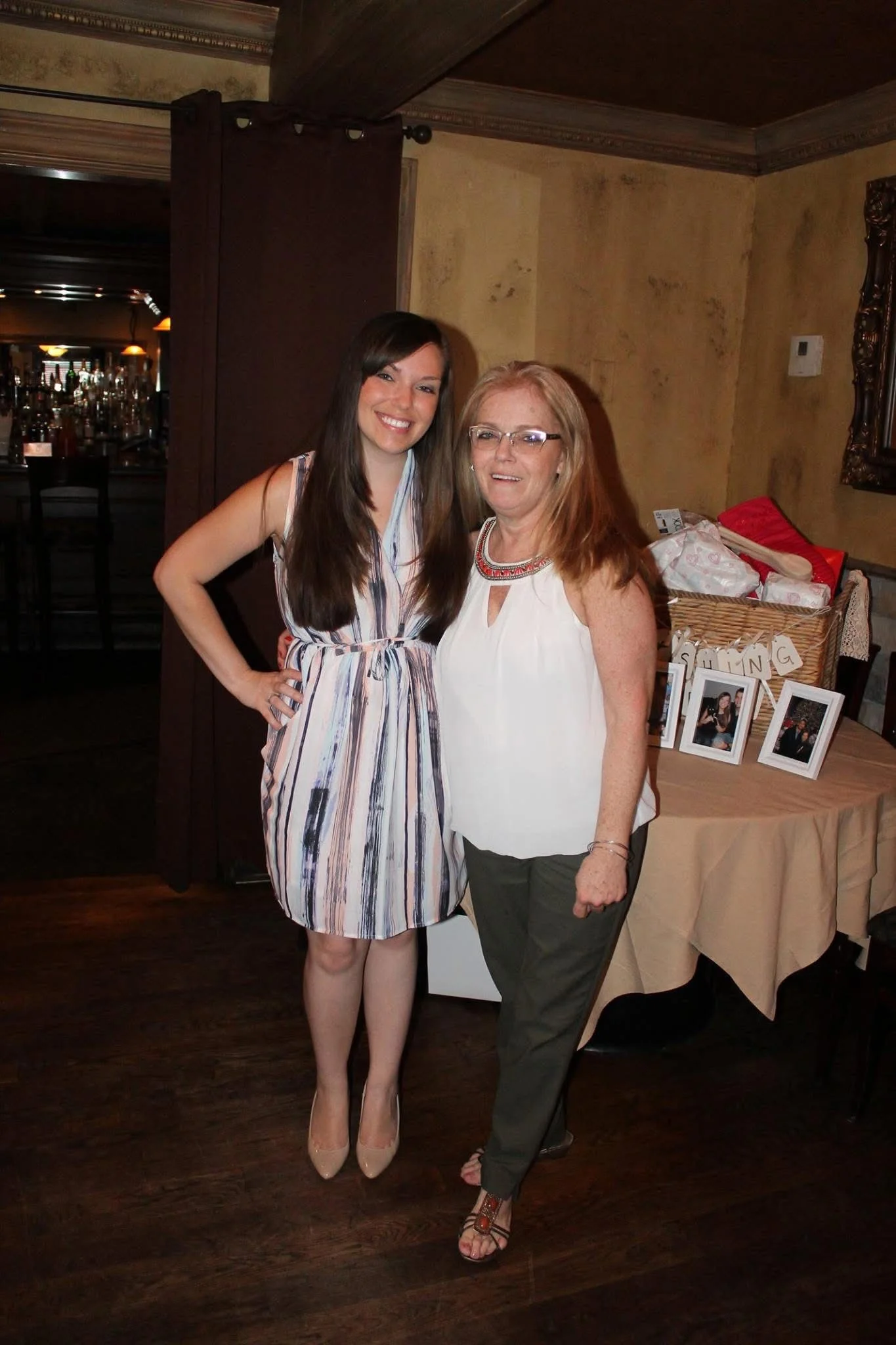 Two women standing together in a cozy restaurant or bar, smiling at the camera. One woman is dressed in a striped, sleeveless dress and beige heels, the other is in a white sleeveless top, black pants, and sandals, with glasses and long reddish hair.