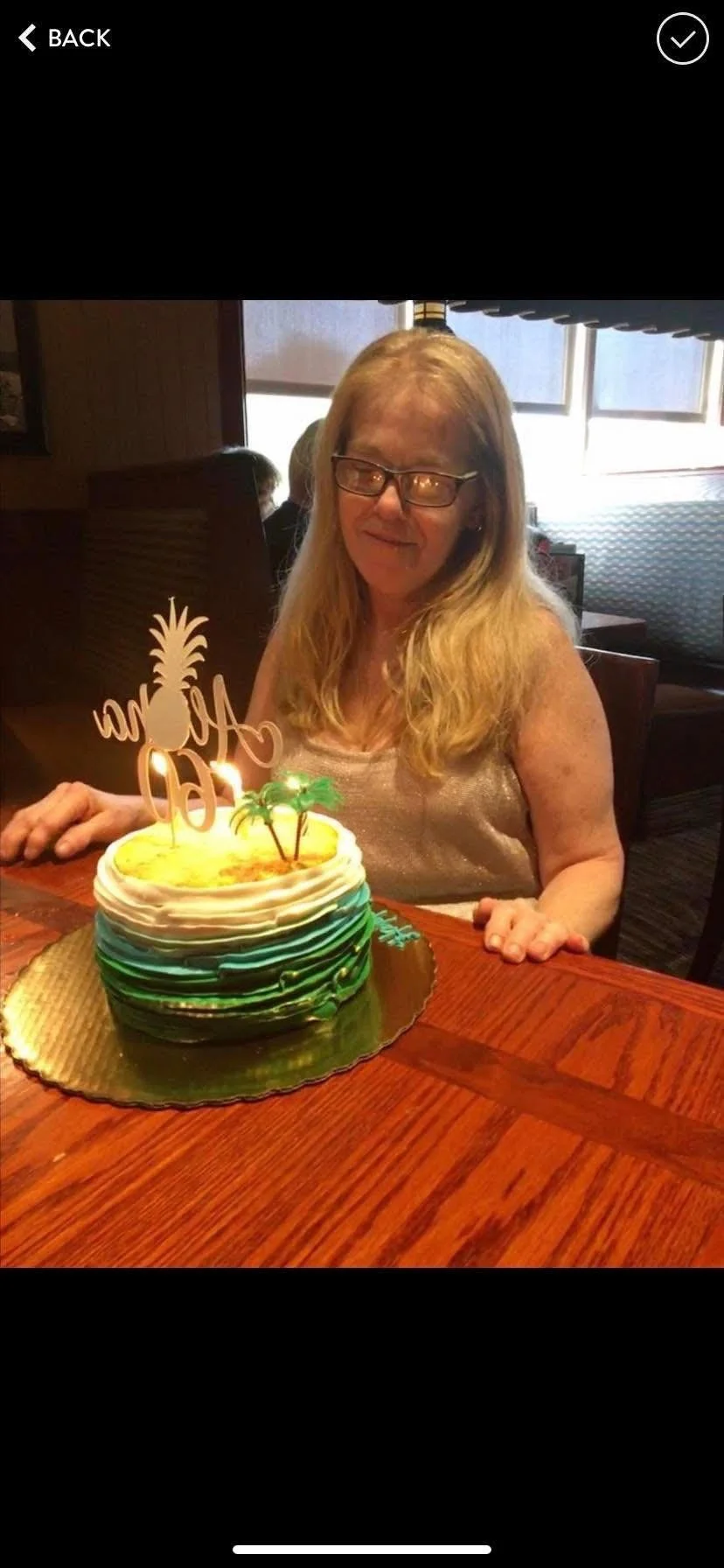 A woman with long blonde hair and glasses sitting at a table with a colorful birthday cake with candles on top.