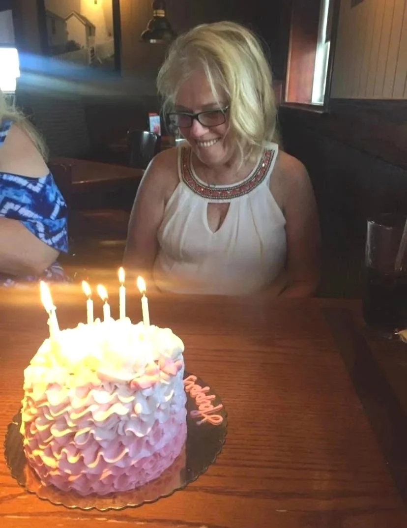A woman with blonde hair and glasses smiling at a birthday cake with lit candles on a wooden table in a restaurant.