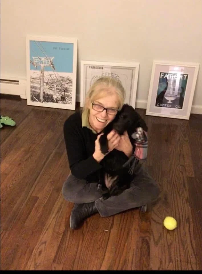 A woman sitting on the wooden floor holding a black dog, with framed artwork behind her and a yellow tennis ball on the floor in front of her.