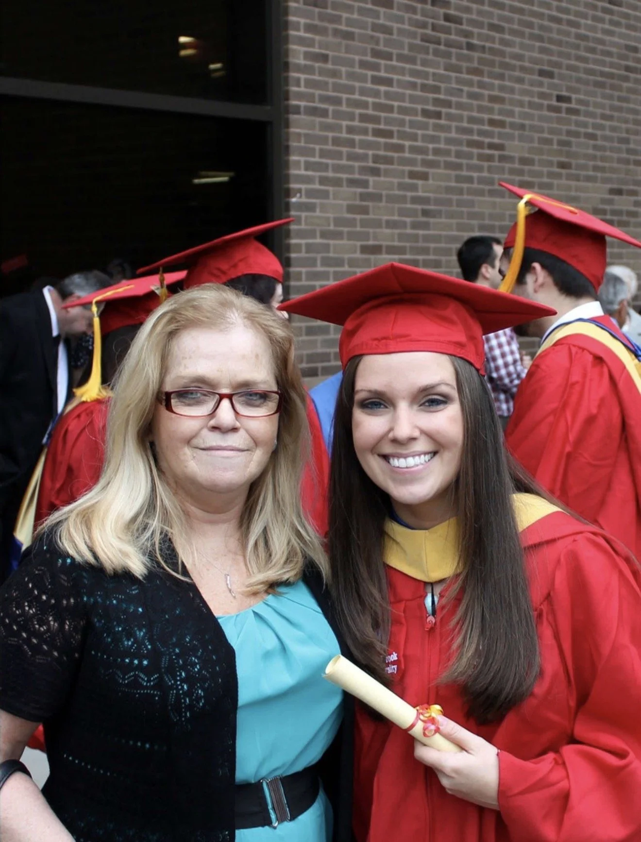 Two women, one older with blonde hair and glasses in a black dress, and one younger with long brown hair and a red graduation gown with cap, posed for a photo at a graduation ceremony.
