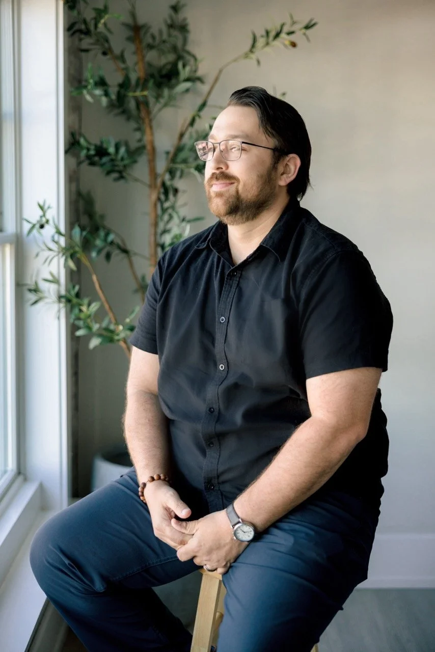 A man with glasses, beard, and dark hair sitting on a stool near a window with sunlight, holding his hands together in his lap, wearing a black short-sleeve shirt, navy pants, a watch, and a beaded bracelet, with a plant in the background.