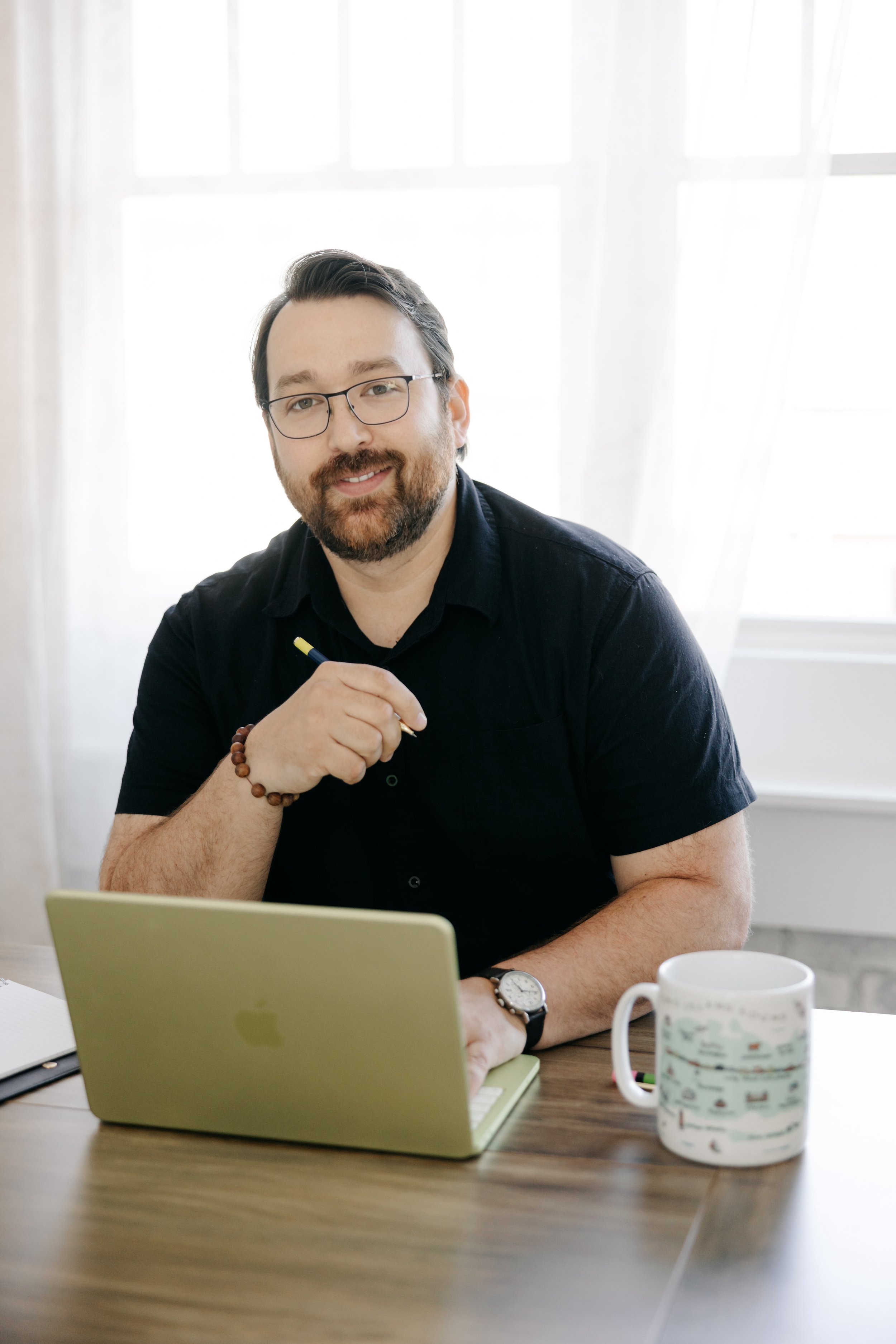 Man with dark hair, glasses, and a beard sitting at a desk with a yellow laptop and a white coffee mug, smiling and holding a pen, in a bright room with white curtains.