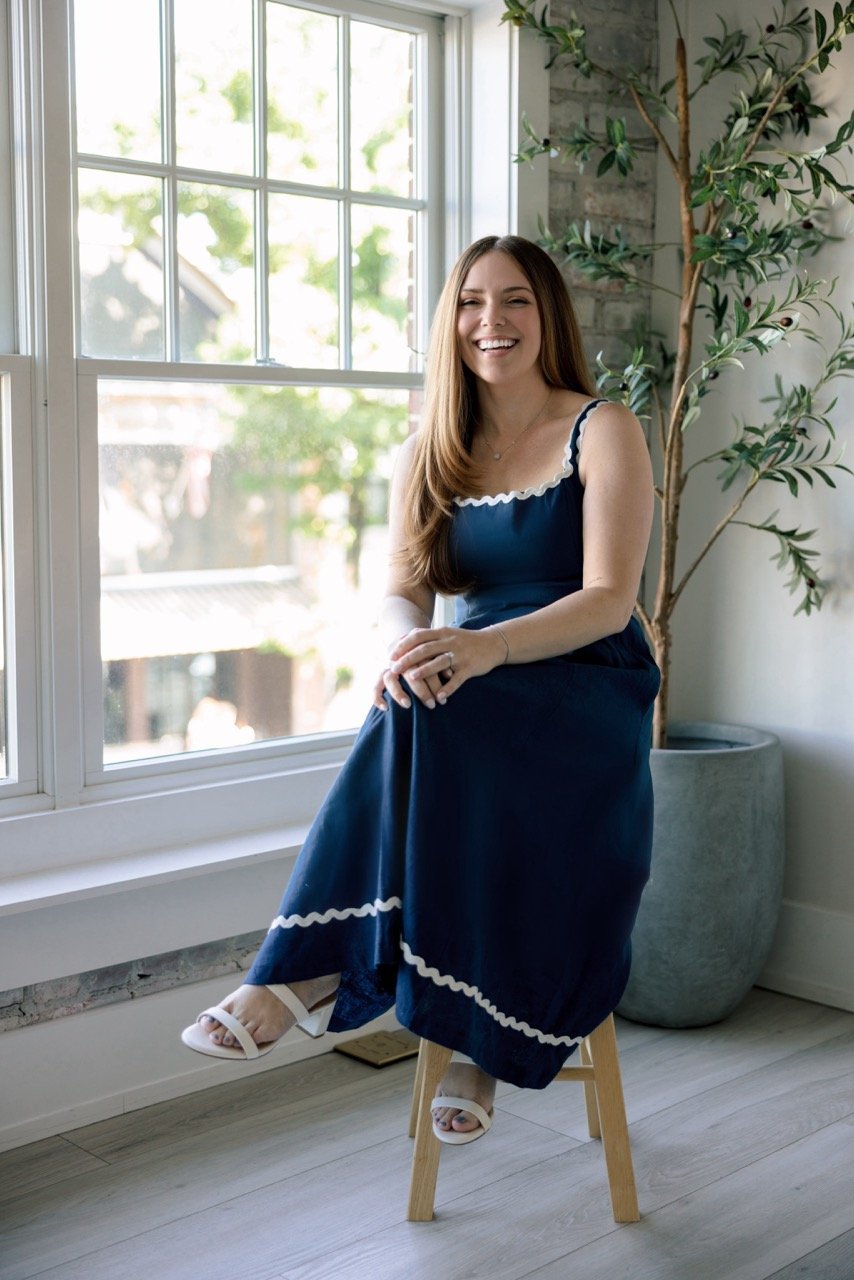 A woman with long brown hair and a blue dress with white accents, white heels, smiling and sitting on a wooden stool by a large window with greenery outside and a potted plant nearby.