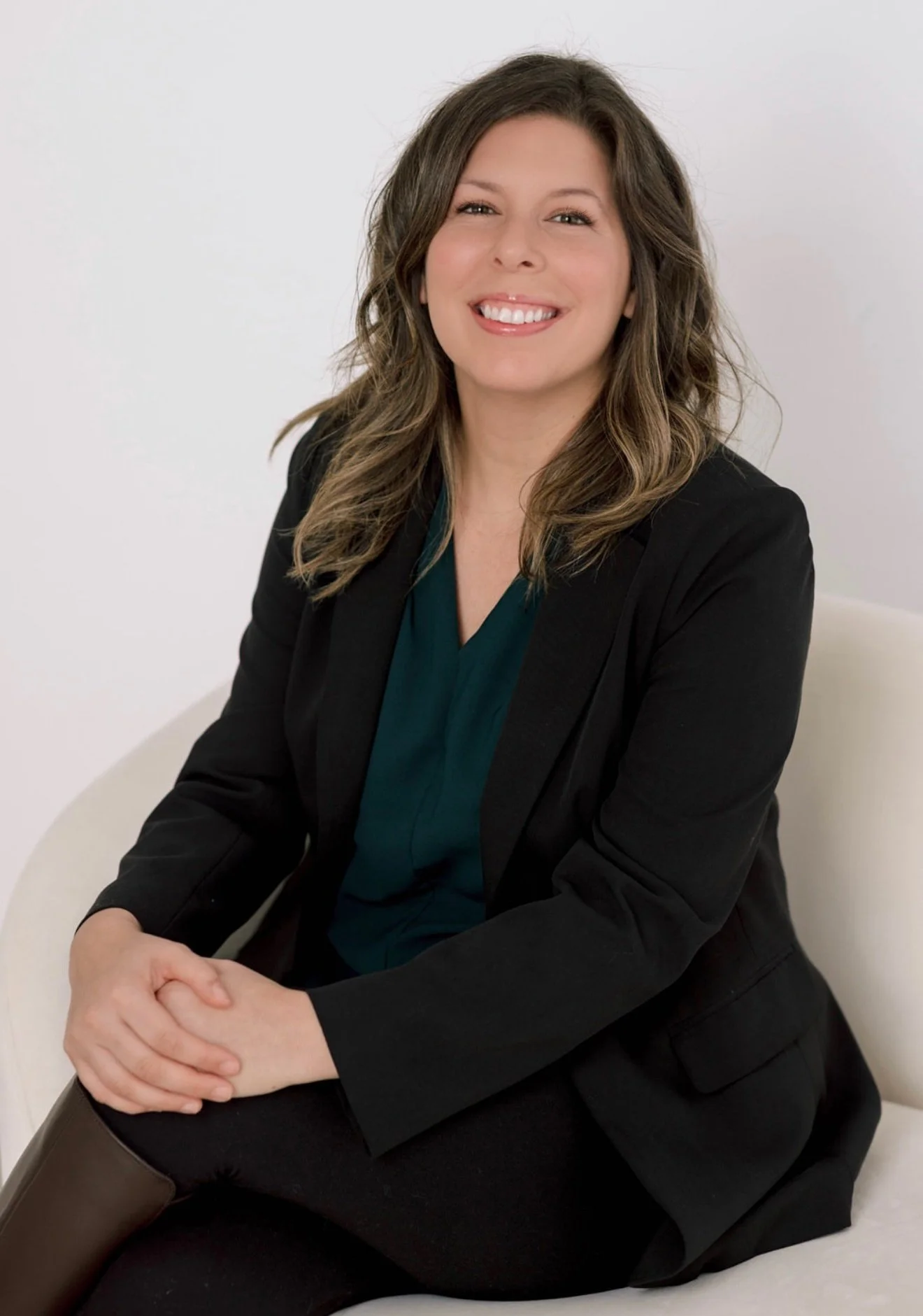 A woman with wavy brown hair, wearing a black blazer and teal top, sitting on a white chair with a plain white background, smiling.
