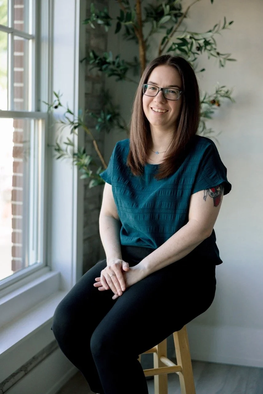 A smiling woman with long brown hair, glasses, and a tattoo on her left arm, sitting on a wooden stool near a large window with sunlight coming in, and a green plant in the background.
