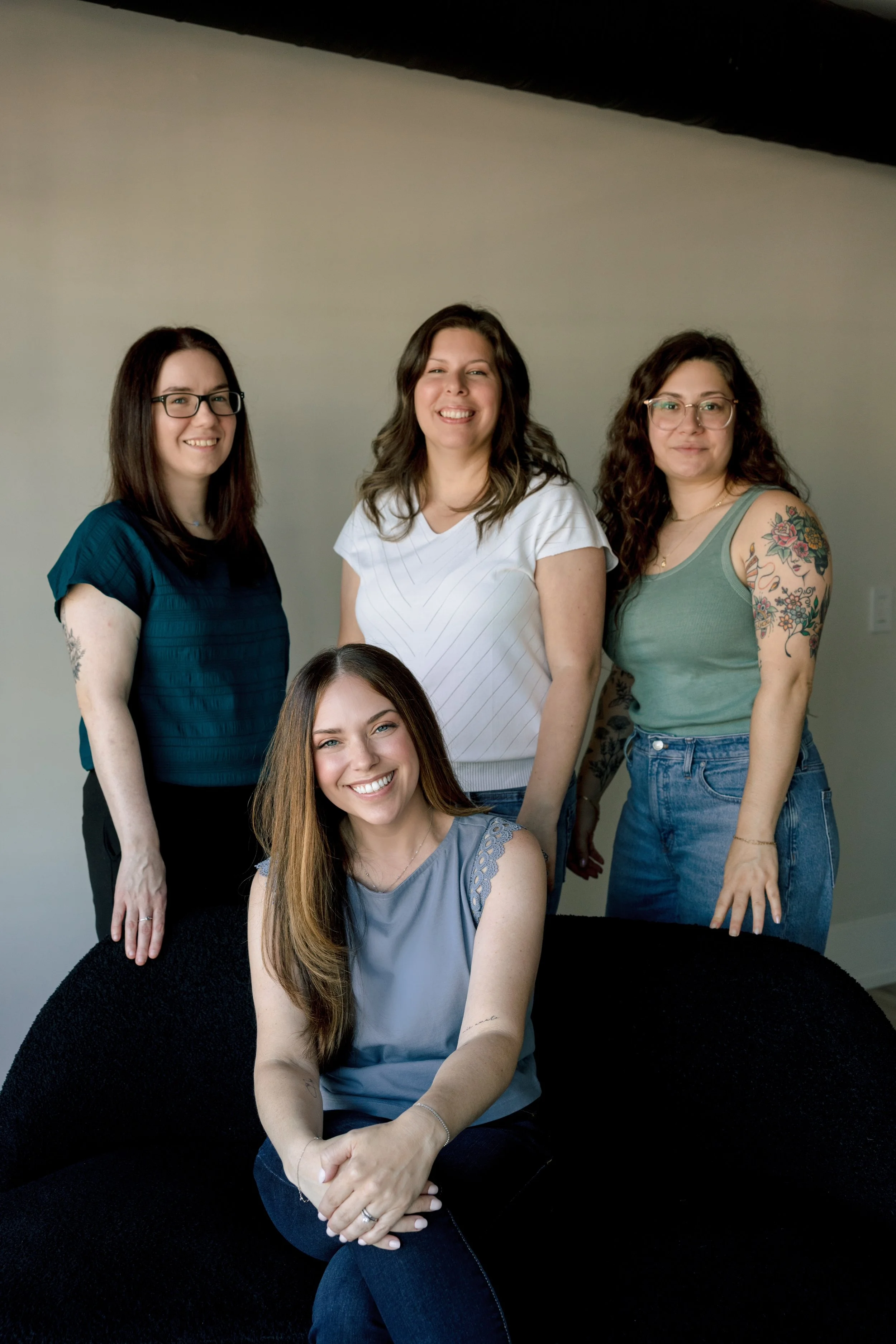 A group of five women in casual attire posing against a plain gray wall, with one woman sitting in front, smiling and the others standing behind her.