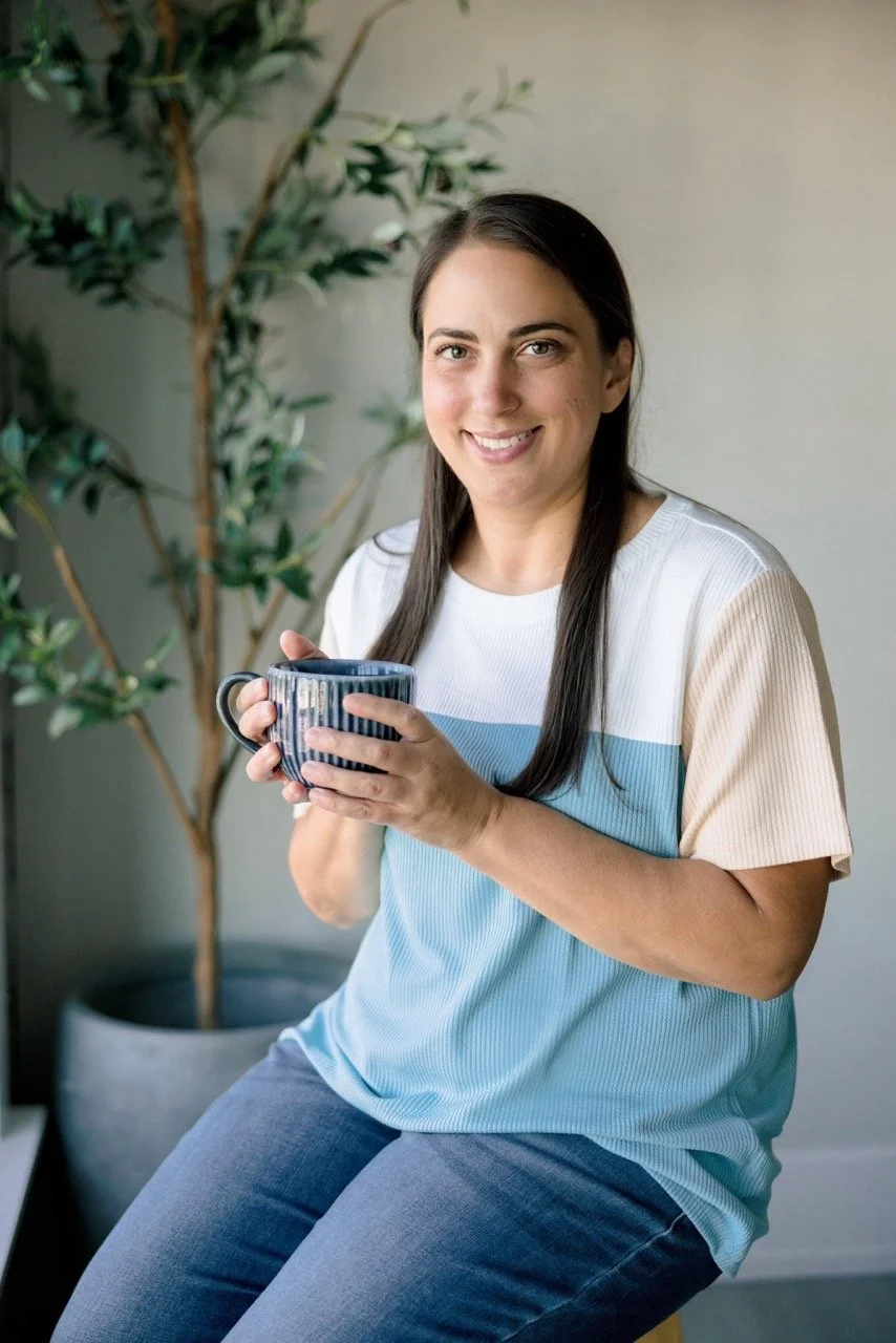 A woman sitting and smiling while holding a blue mug with black stripes, in a room with a large green plant in the background.