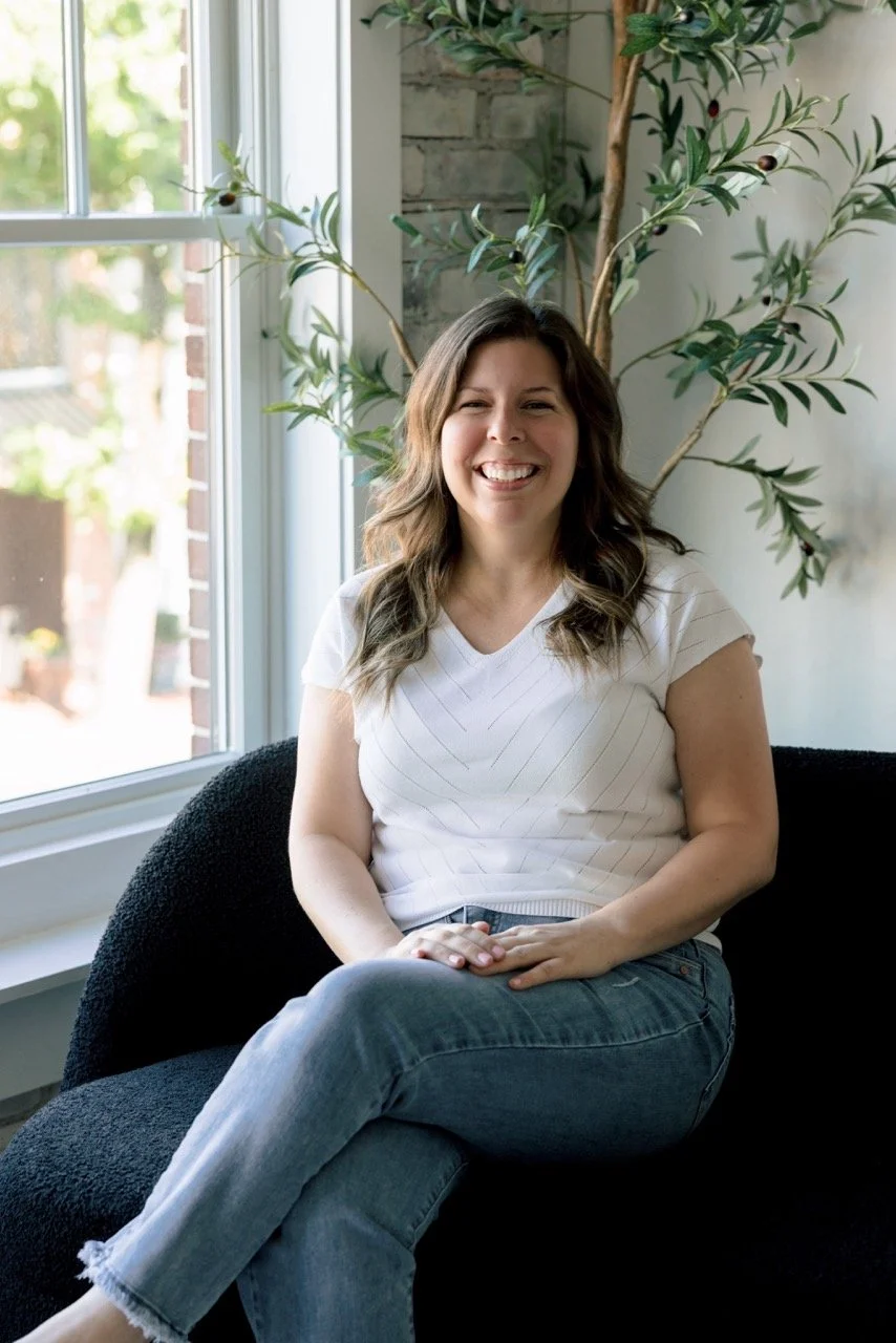 A woman with wavy brown hair sitting on a black couch near a window with sunlight, smiling, with a large plant with olive-like leaves behind her.