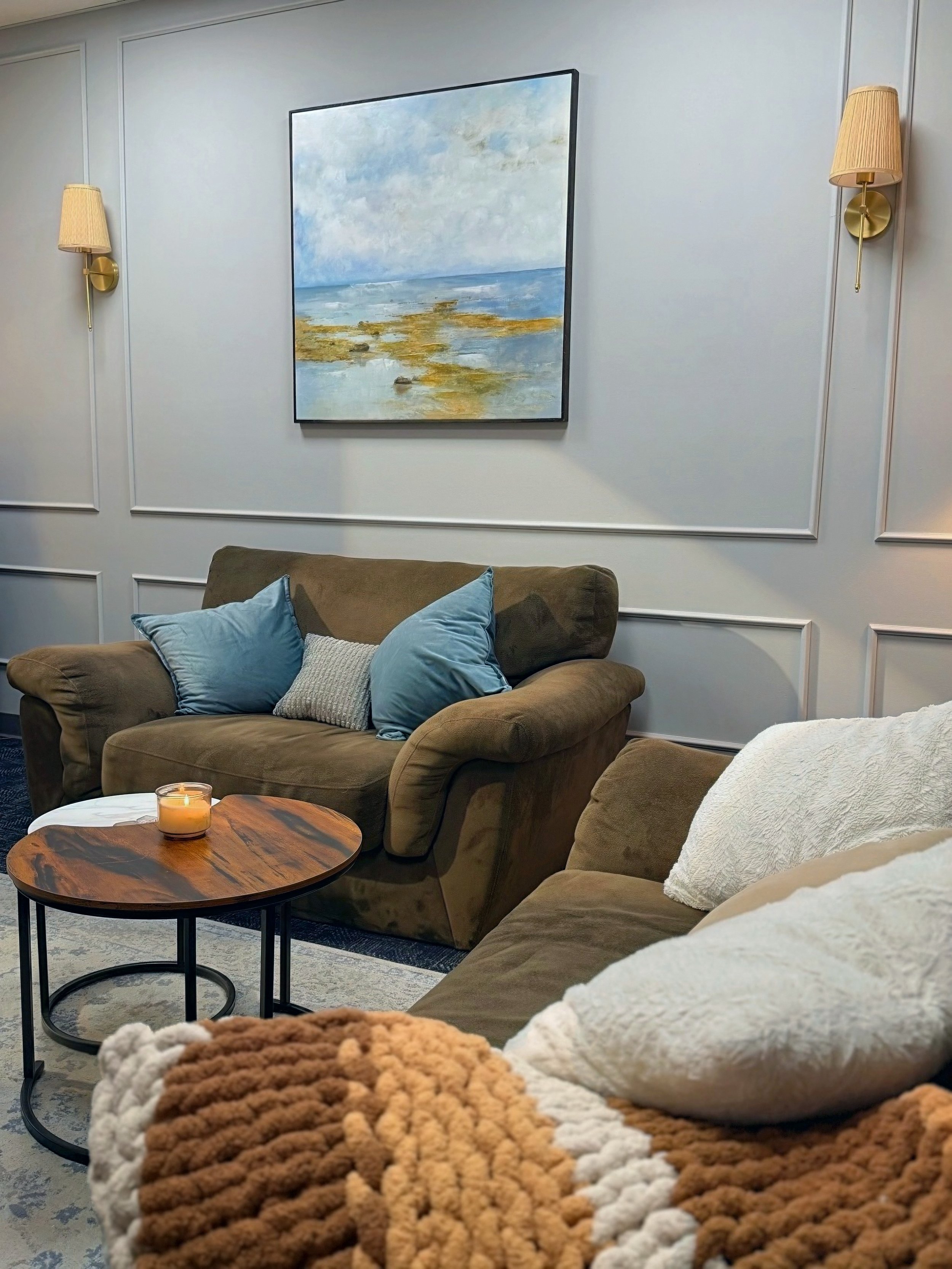 Living room with brown sofas, light blue and white pillows, a round wooden coffee table with a candle, wall-mounted lamps, and a framed painting of a seascape with clouds and water.