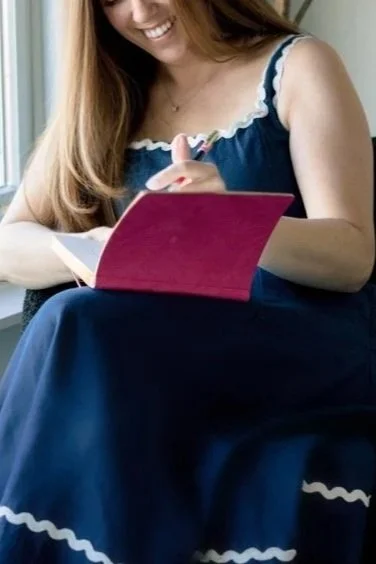 A woman sitting by a window, smiling, writing in a pink notebook.