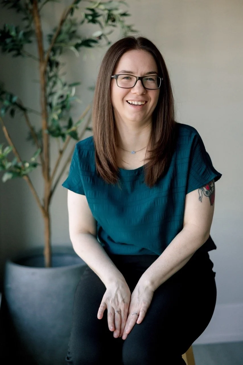A woman with long brown hair wearing glasses, a turquoise blouse, and black pants, sitting indoors with a large potted plant in the background, smiling at the camera.