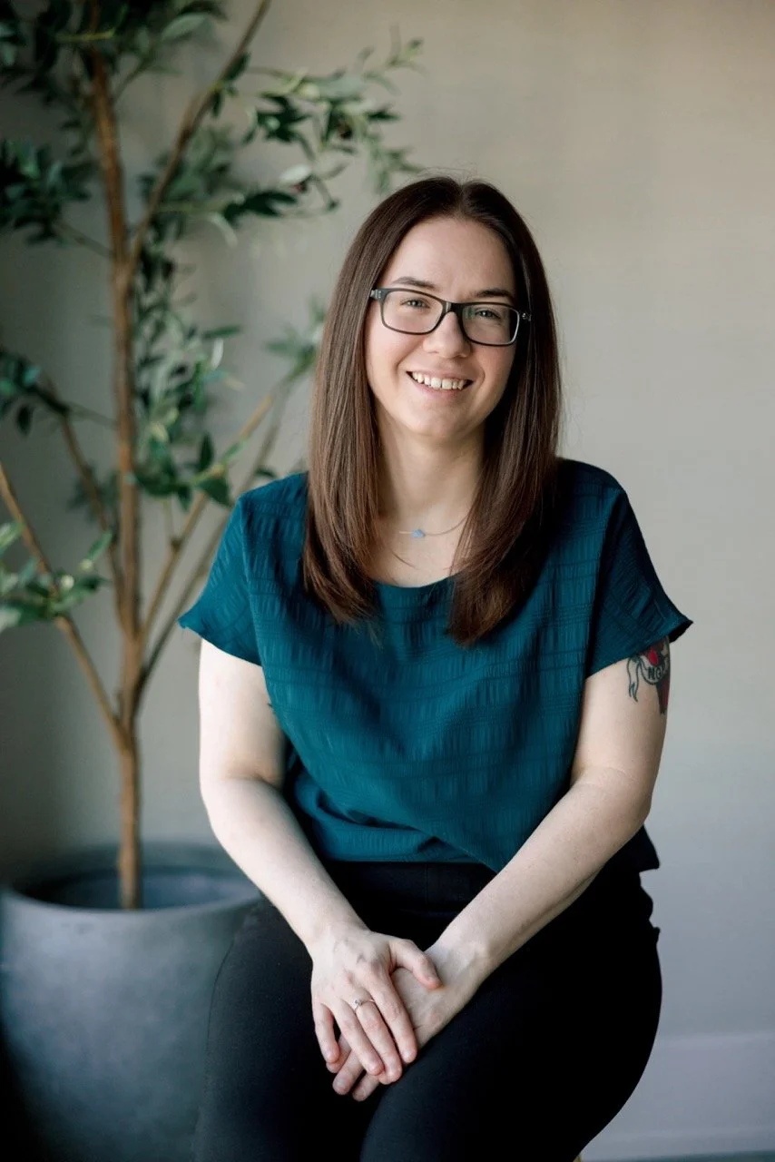 A smiling woman with glasses and long brown hair, wearing a teal blouse, sitting in front of a home plant.