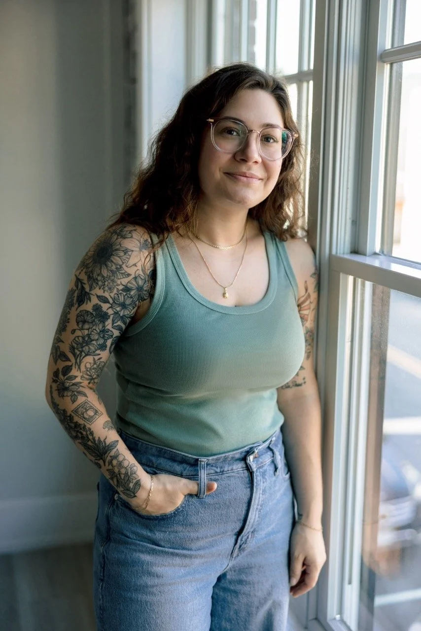 A young woman with wavy brown hair, glasses, and tattoos on her arms, wearing a green tank top and jeans, standing by a window.