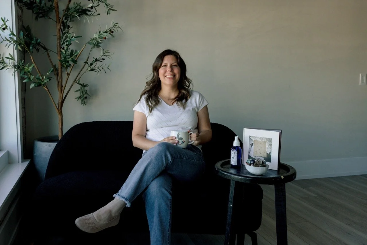 A young woman sitting on a black sofa, smiling and holding a white mug, in a cozy living room with a tall potted plant, a small black side table with a framed picture, a bottle of hand sanitizer, and a bowl of decorative stones.
