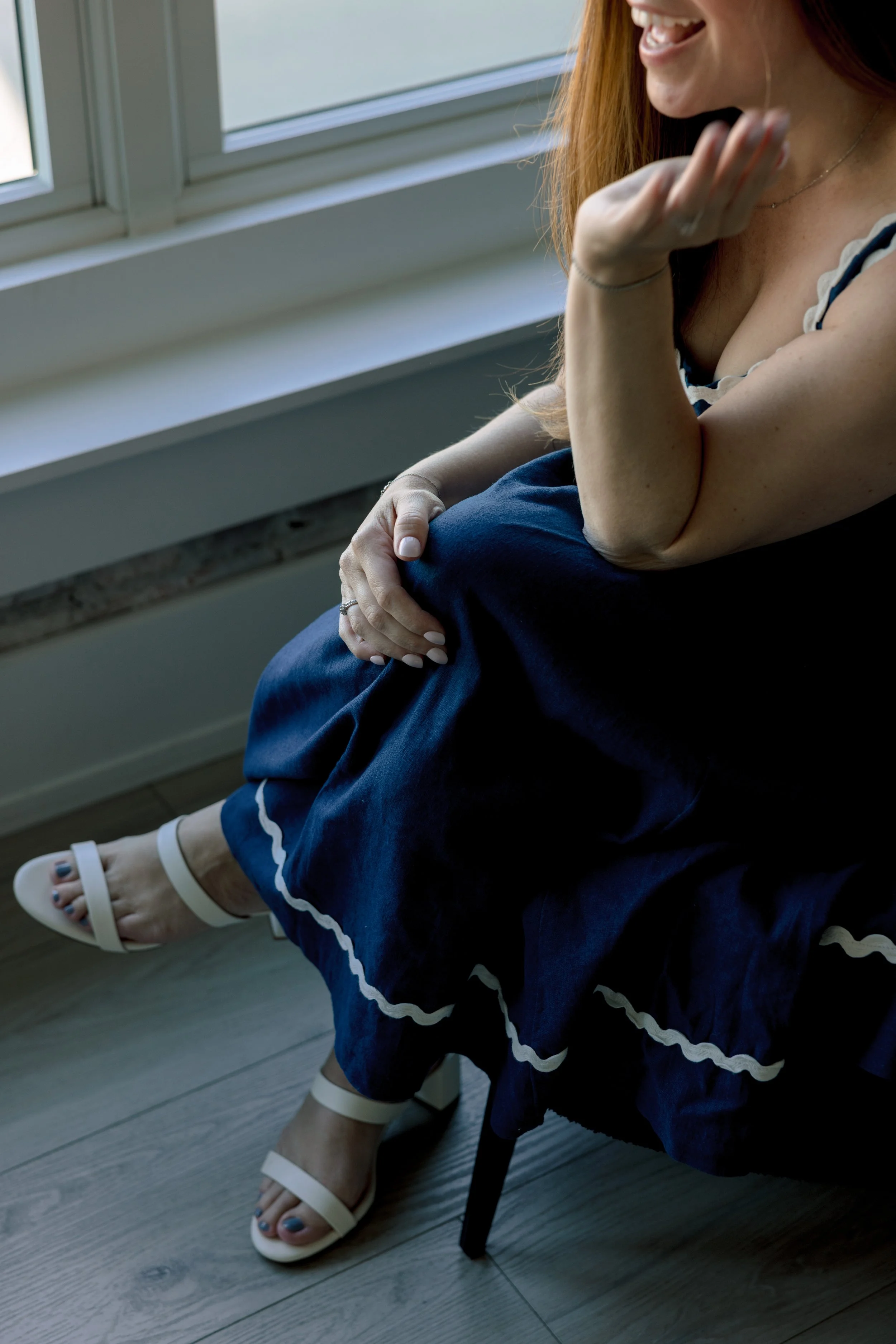 A woman with red hair sitting near a window, smiling and touching her face. She is wearing a blue dress with white stripes, white sandals, and has her nails painted blue.