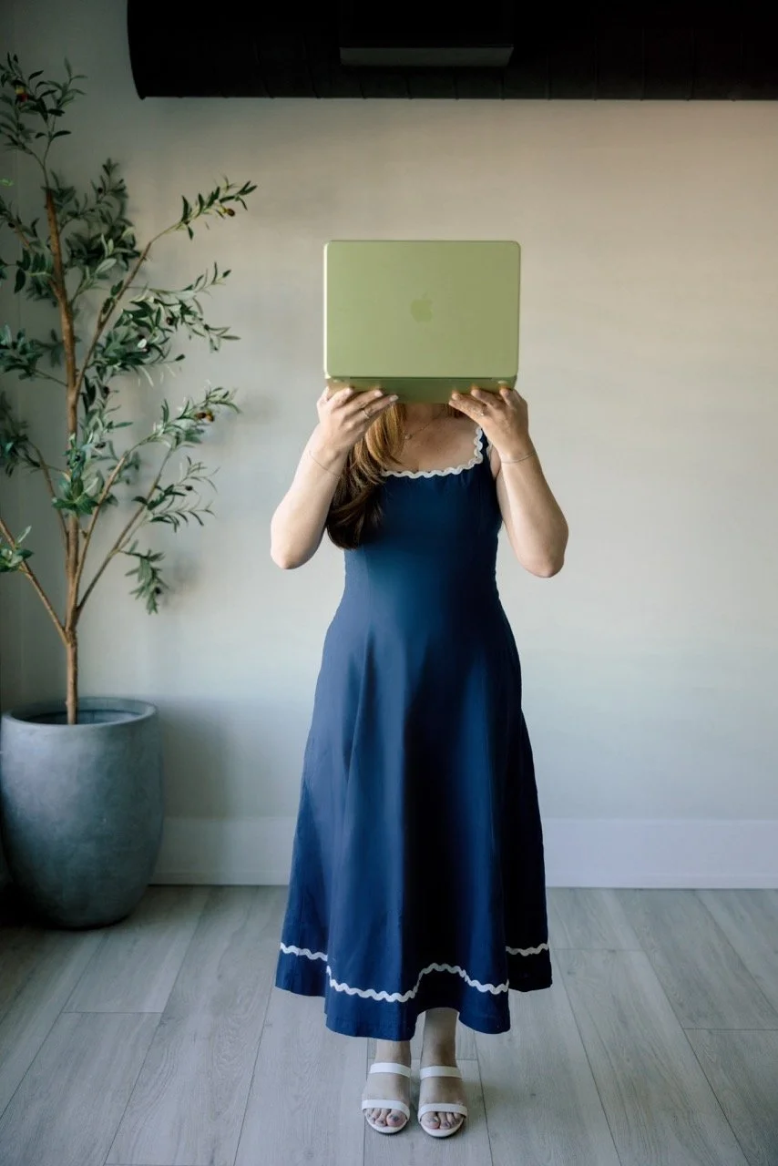 A woman in a blue dress with white trim is standing indoors, holding a light green laptop in front of her face. There is a potted plant with green leaves to her left and a plain light-colored wall behind her. She is wearing white sandals and standing on a wood floor.
