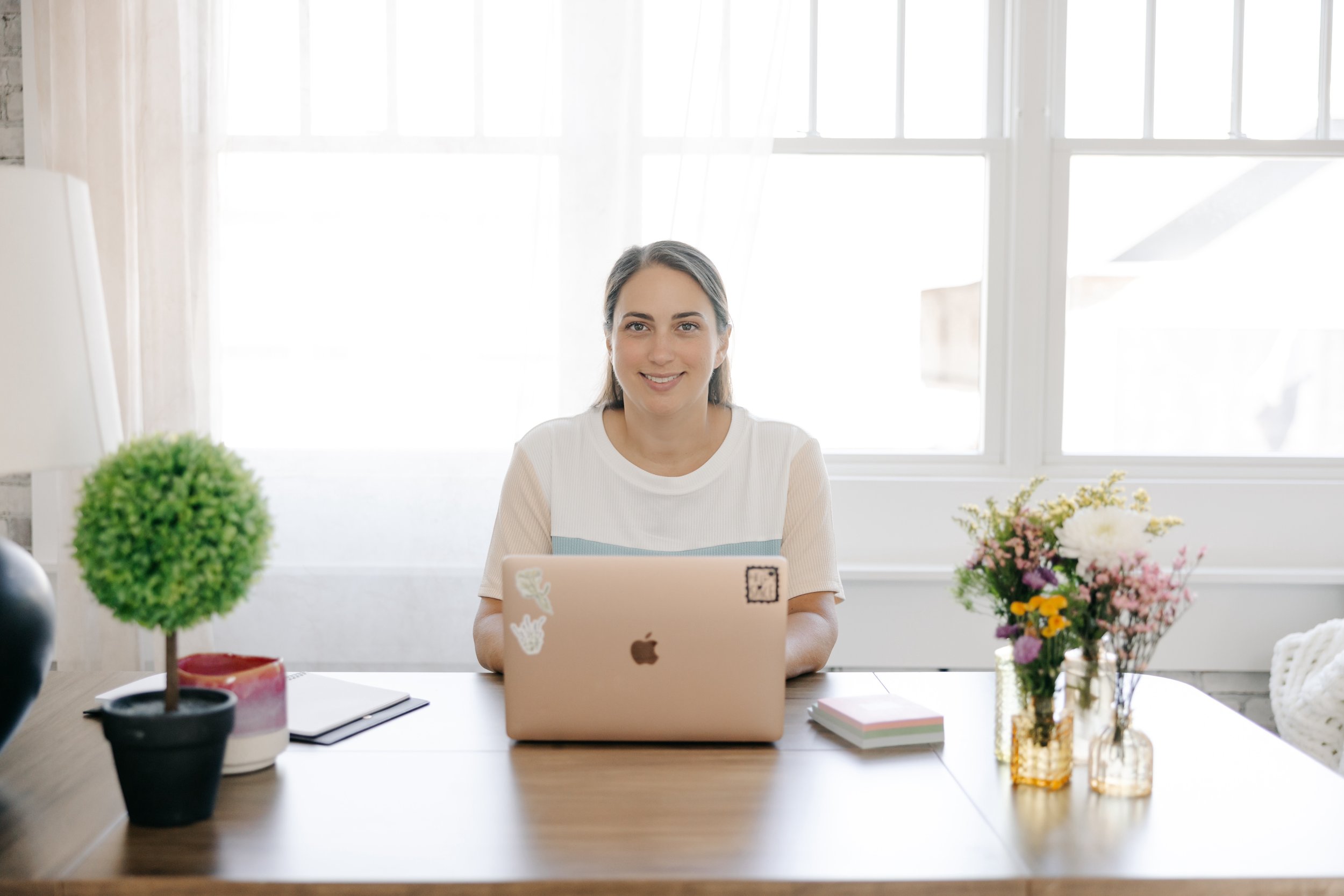 A woman sitting at a table with a laptop, smiling, in a bright room with large windows, surrounded by colorful flower arrangements and decor.
