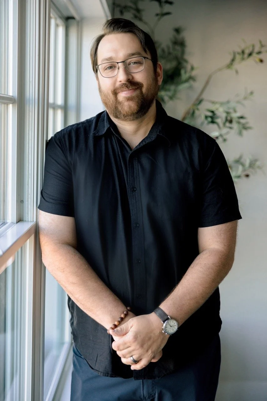 A man with glasses and a beard, wearing a black short-sleeved shirt, standing near a window with natural light, with a plant in the background.