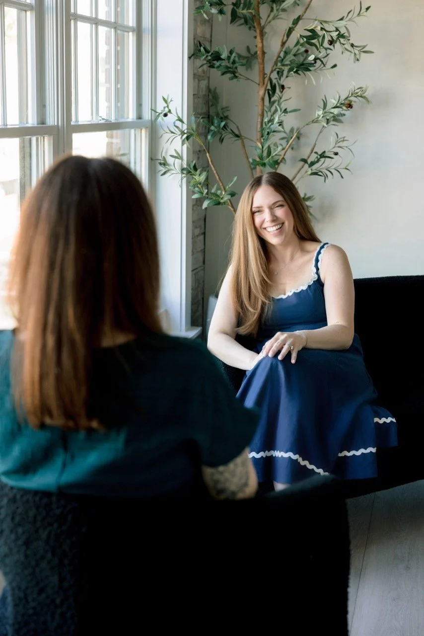 Two women having a conversation near a large window with natural light, one woman with long brown hair in a blue dress smiling, the other with red hair in a dark outfit.