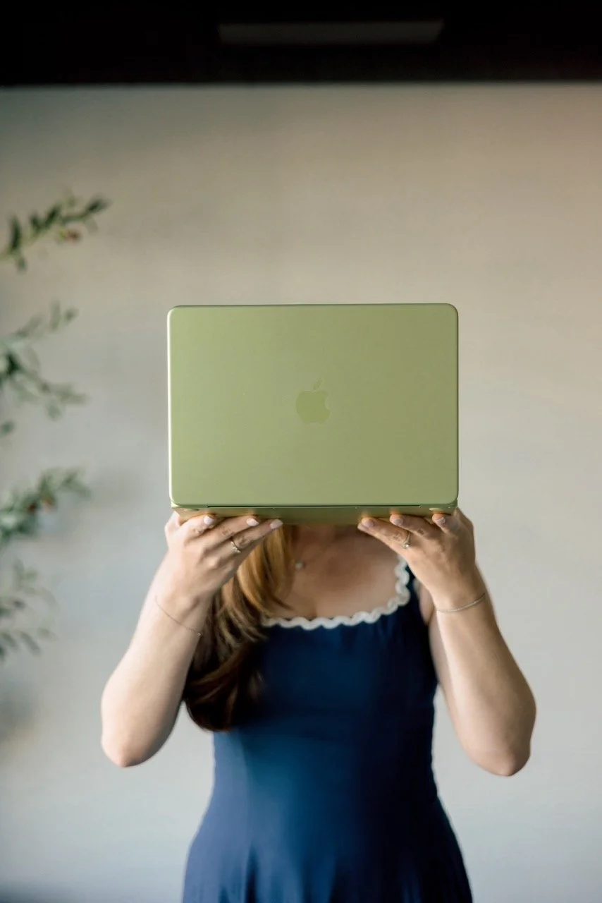 Woman holding a green Apple laptop in front of her face, standing indoors with a blurry plant in the background.