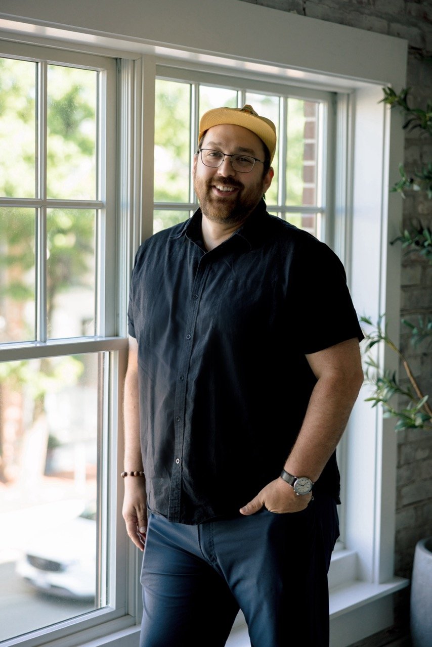 Man with glasses, beard, and yellow cap smiling, standing indoors by a large window with green trees outside, wearing a black shirt, navy pants, watch, and bracelet.