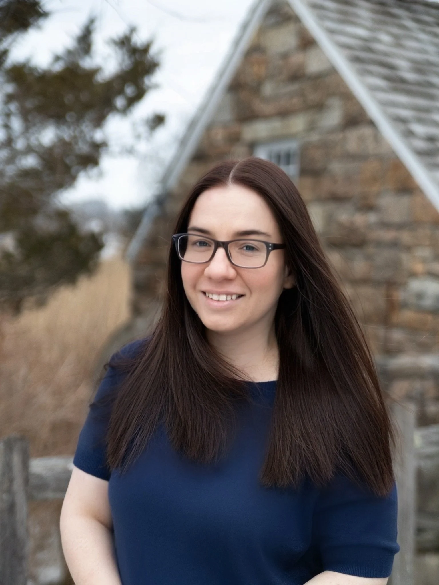 A young woman with long brown hair and glasses smiling outdoors, standing in front of a stone building with trees in the background.