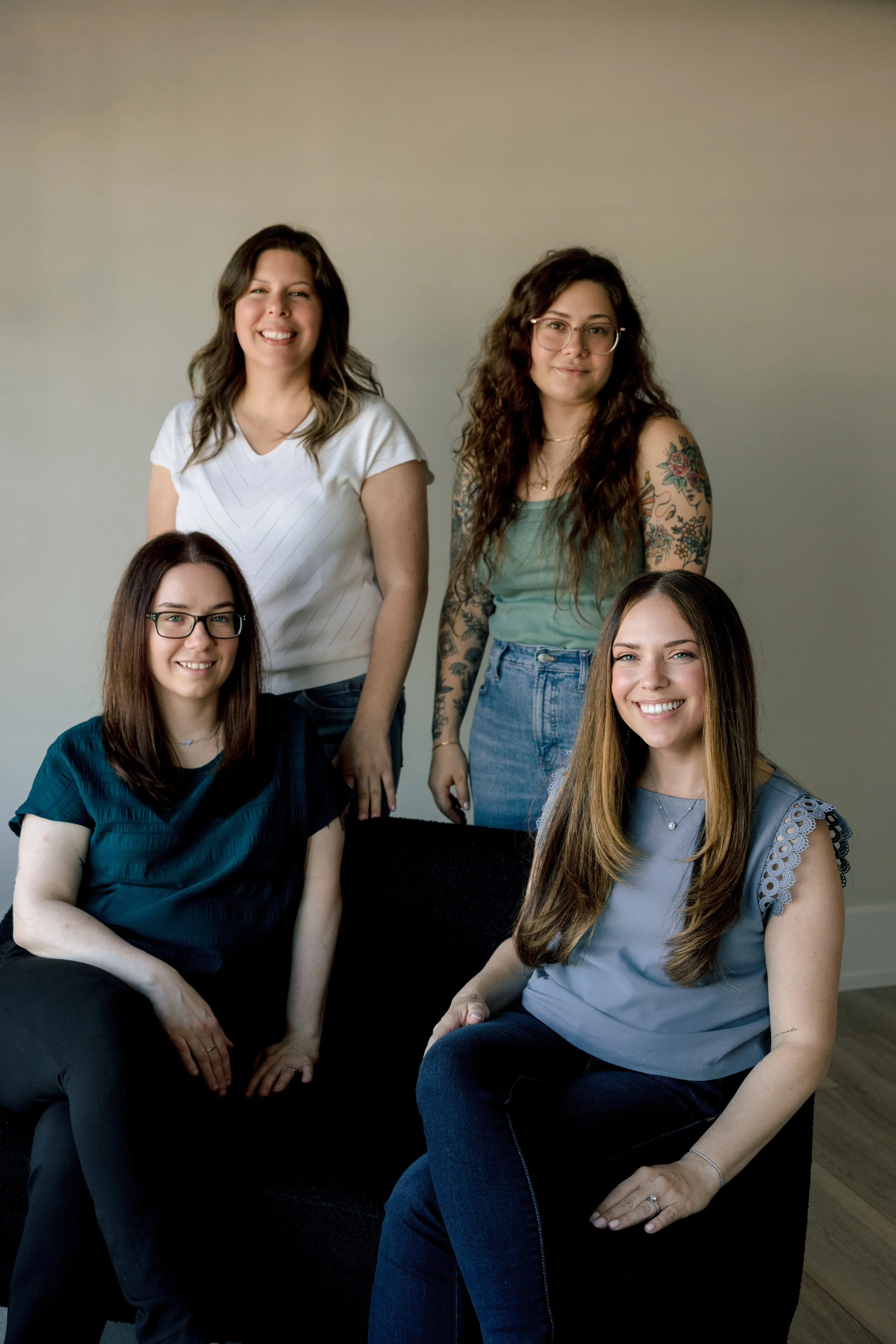 Four women sitting and standing in front of a plain beige wall, smiling at the camera. They have long hair, glasses, and tattoos, wearing casual tops and jeans.