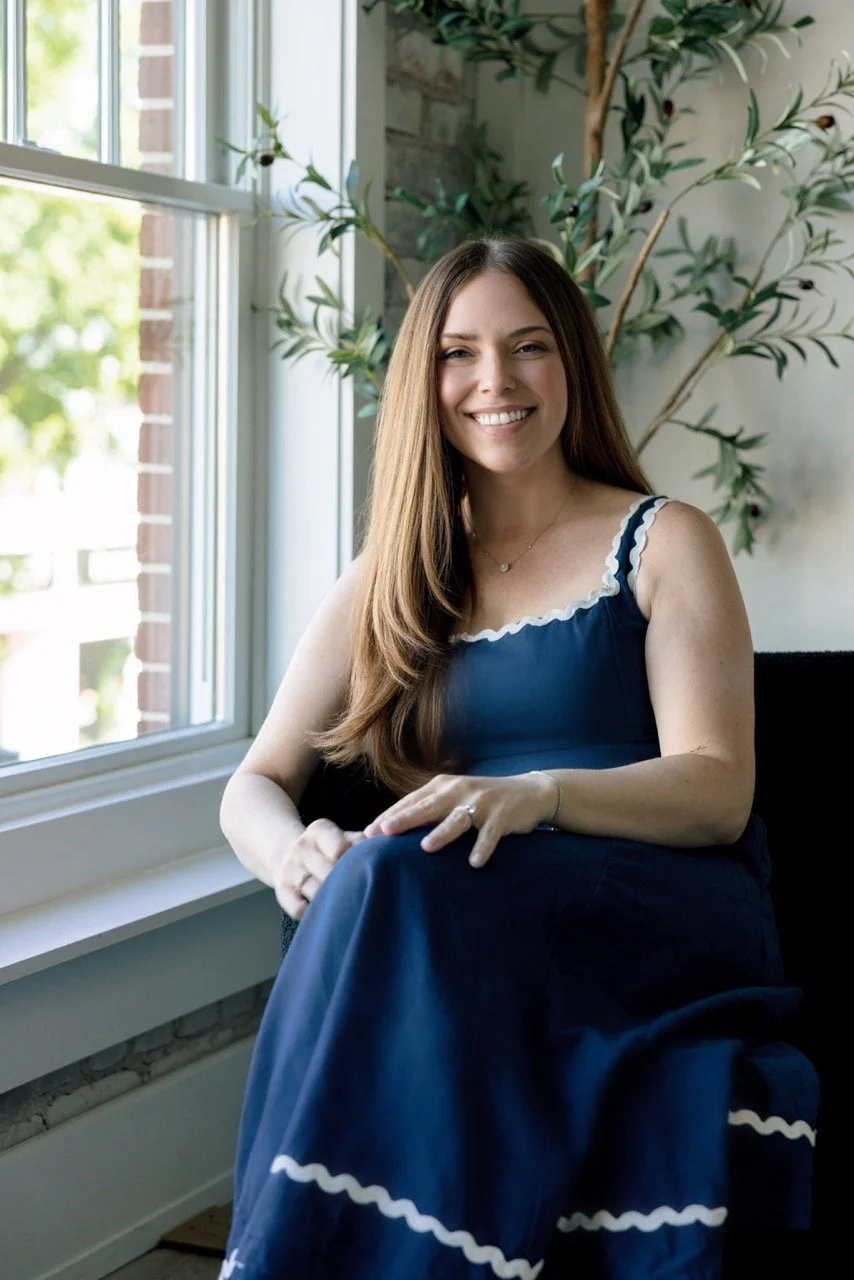 A young woman with long brown hair sitting by a large window, smiling, wearing a navy blue dress with white trim, in a room with a potted plant and brick wall.