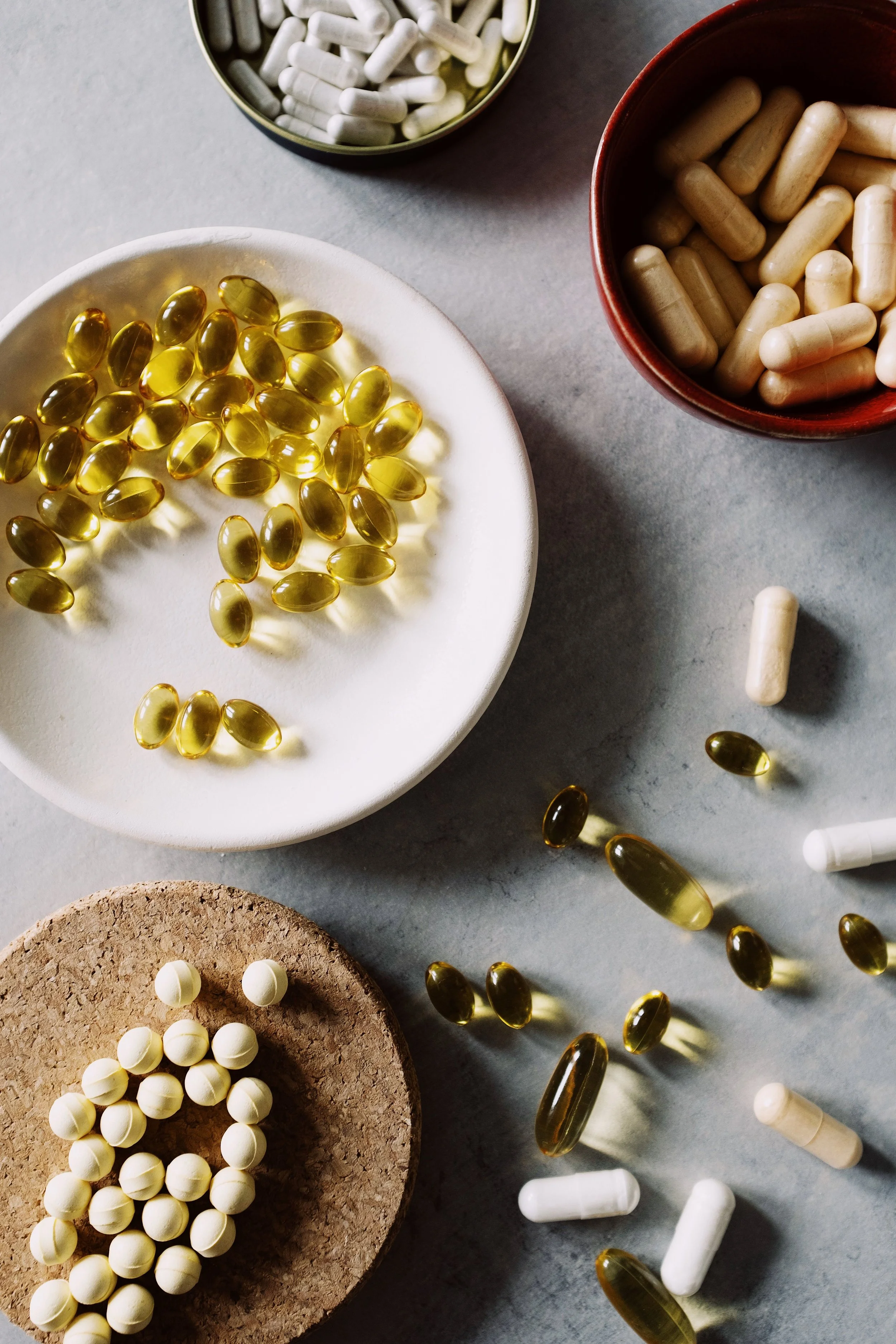 Assorted pills and capsules on bowls, cork, and scattered on a gray surface