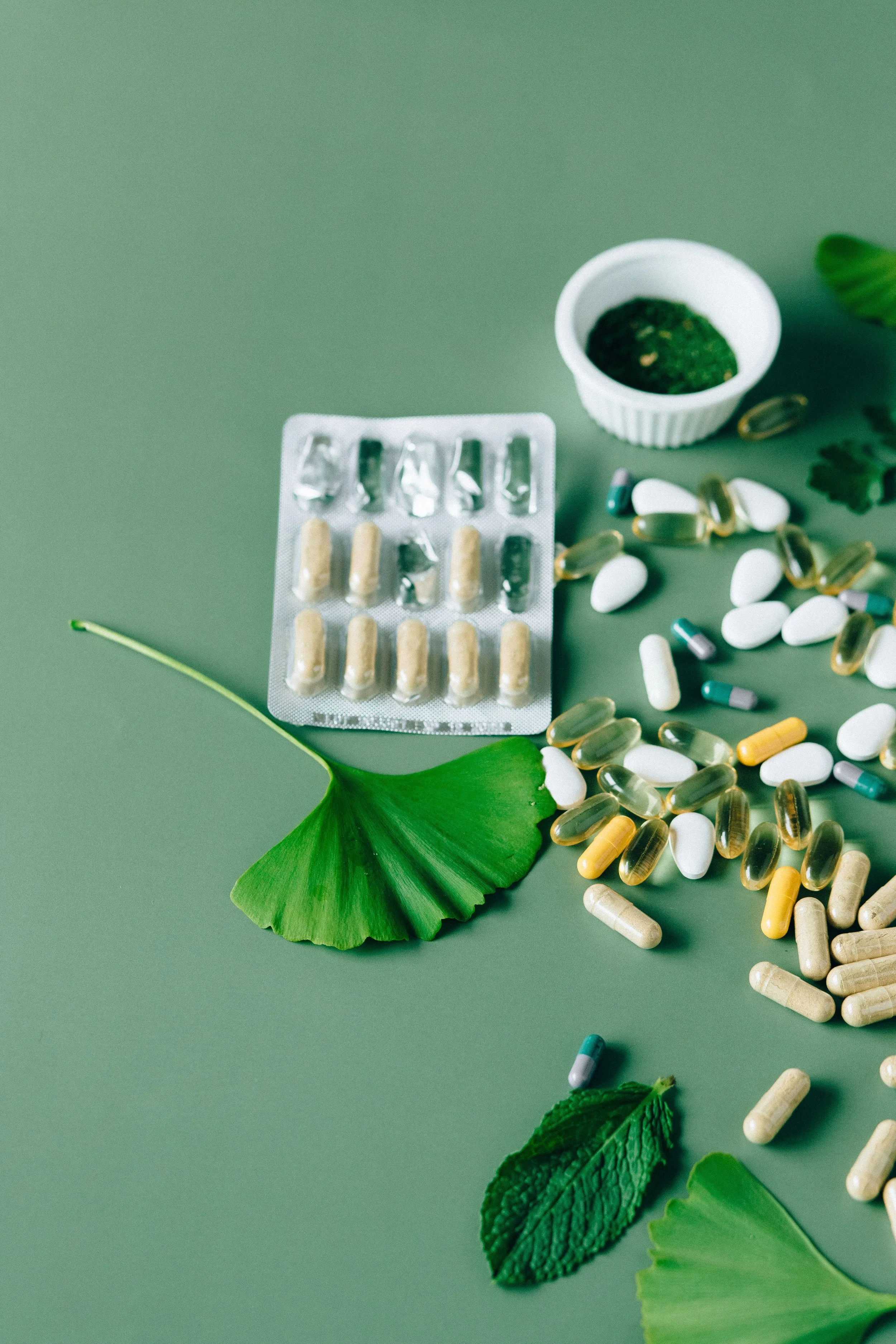 Various pills and capsules scattered on a green surface with green leaves and a small container with a green substance.