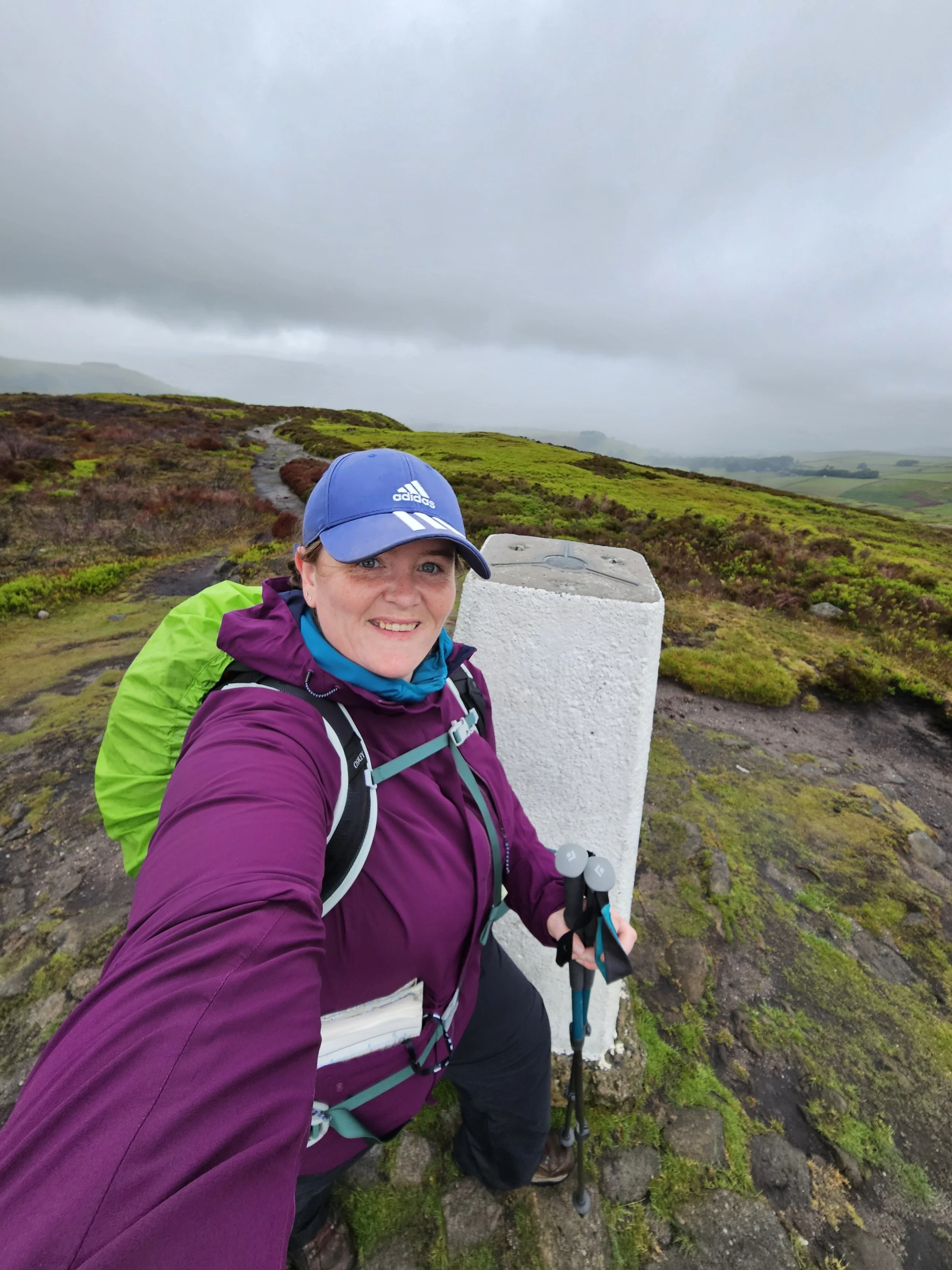A woman in outdoor hiking gear, including a blue cap and purple jacket, taking a selfie at a mountain summit with a trail and scenic landscape in the background. She is holding trekking poles and standing next to a white milestone marker.