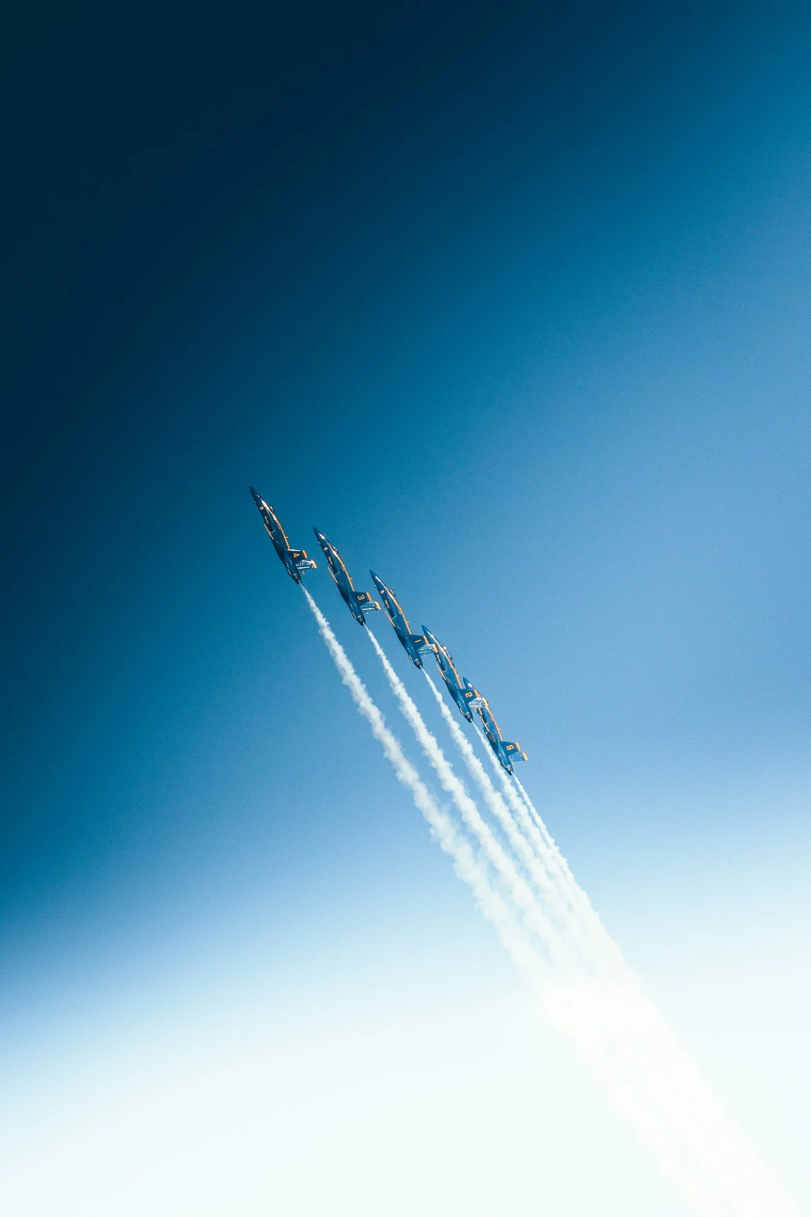 Aerial view of five fighter jets flying in a tight formation, leaving white smoke trails against a bright blue sky.
