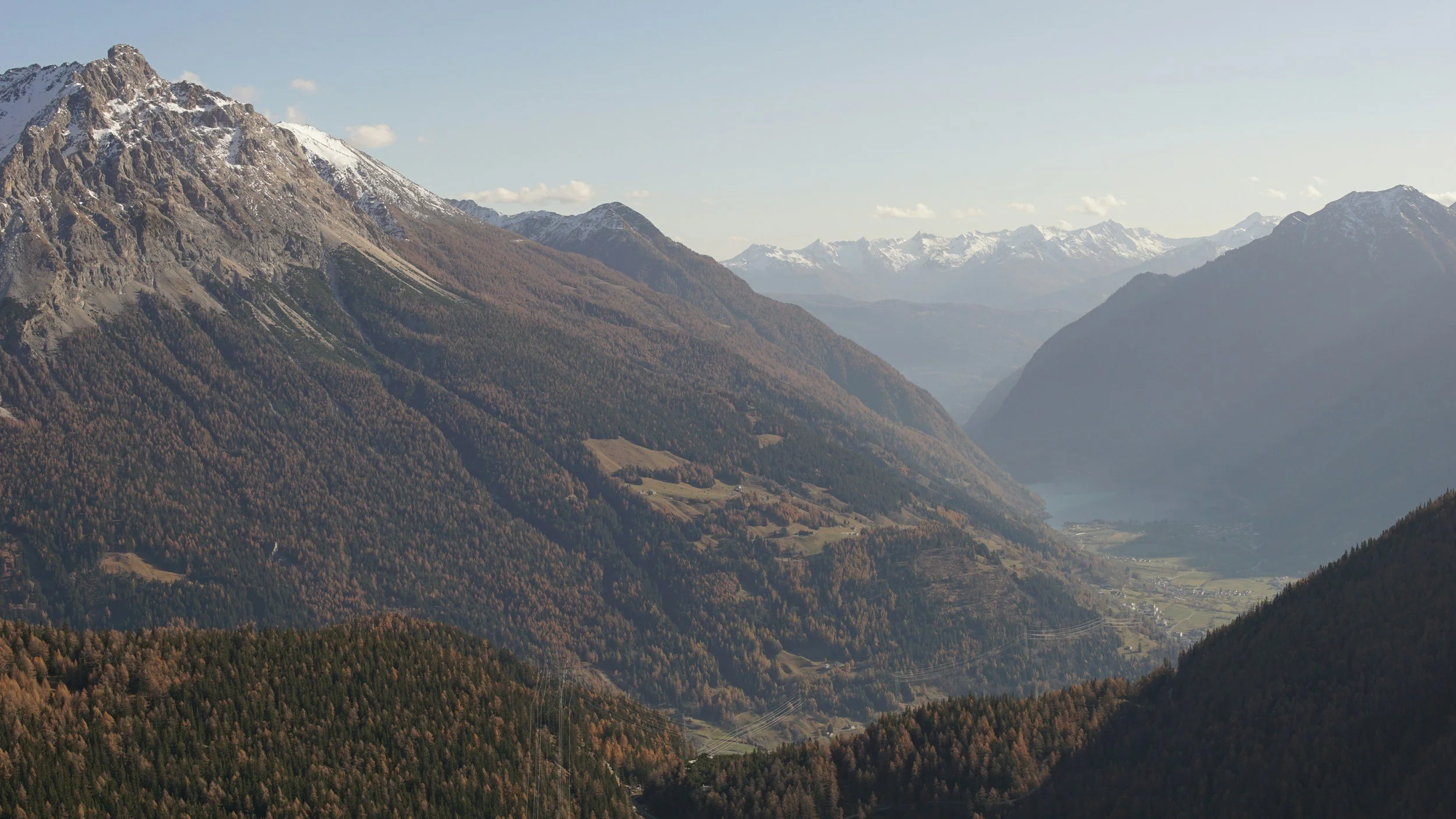 Berglandschaft mit schneebedeckten Gipfeln im Hintergrund und bewaldeten Hängen im Vordergrund, durch eine Valley im Tal fließender Fluss sichtbar.