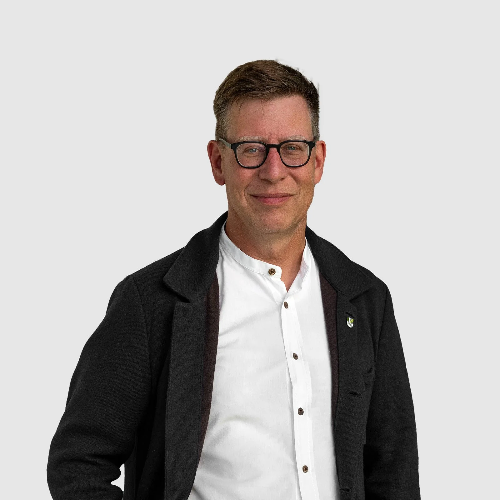 Ein Mann trägt eine dunkle Jacke und eine weiße Hemd, trägt schwarze Brille, lächelt leicht, hat kurzes braunes Haar, steht vor hellem Hintergrund.
