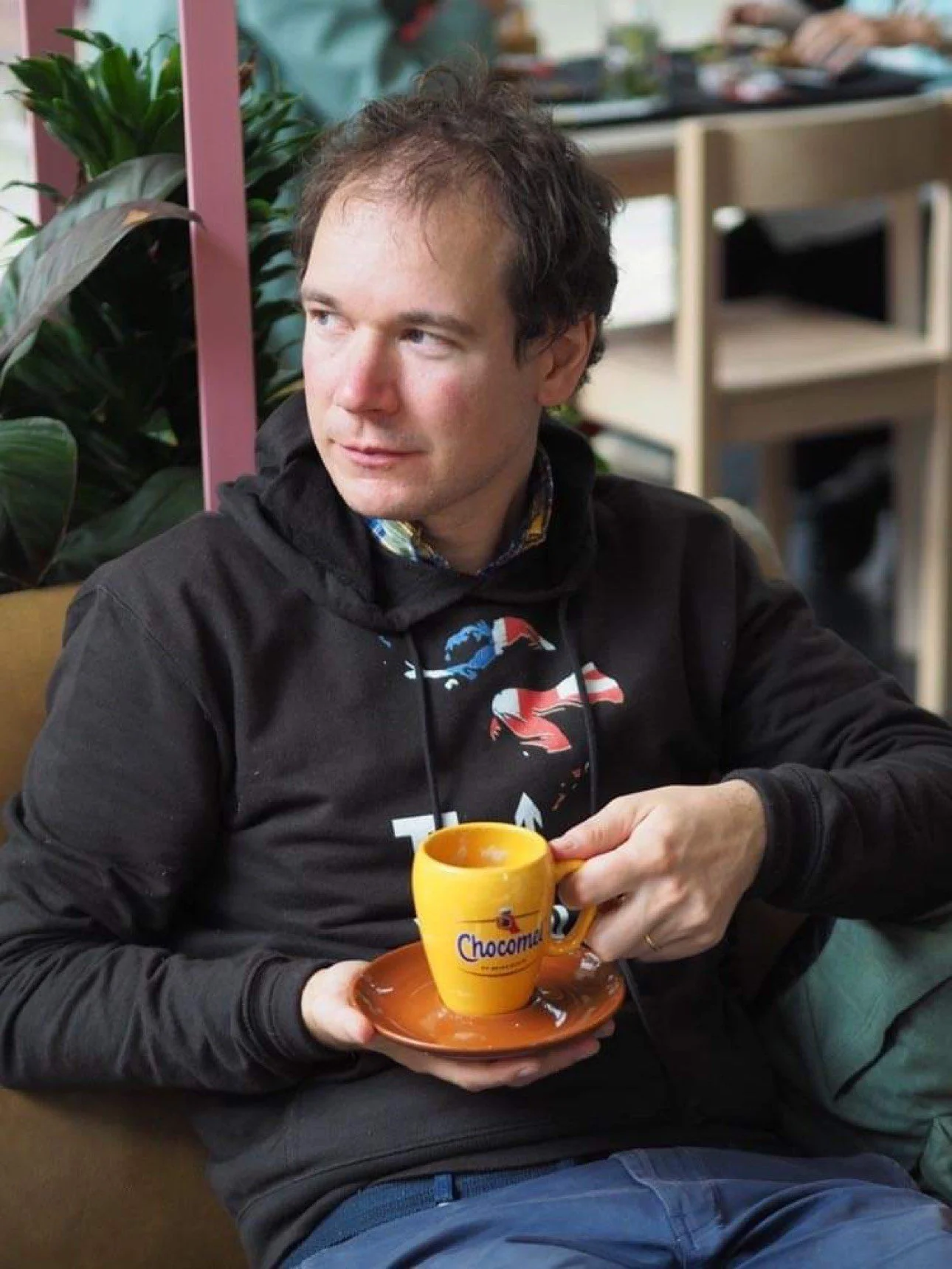 A man sitting on a couch holding a yellow mug on a matching saucer. The man is wearing a black hoodie with a graphic design. In the background, there are green plants and wooden shelves with books and decorative items.