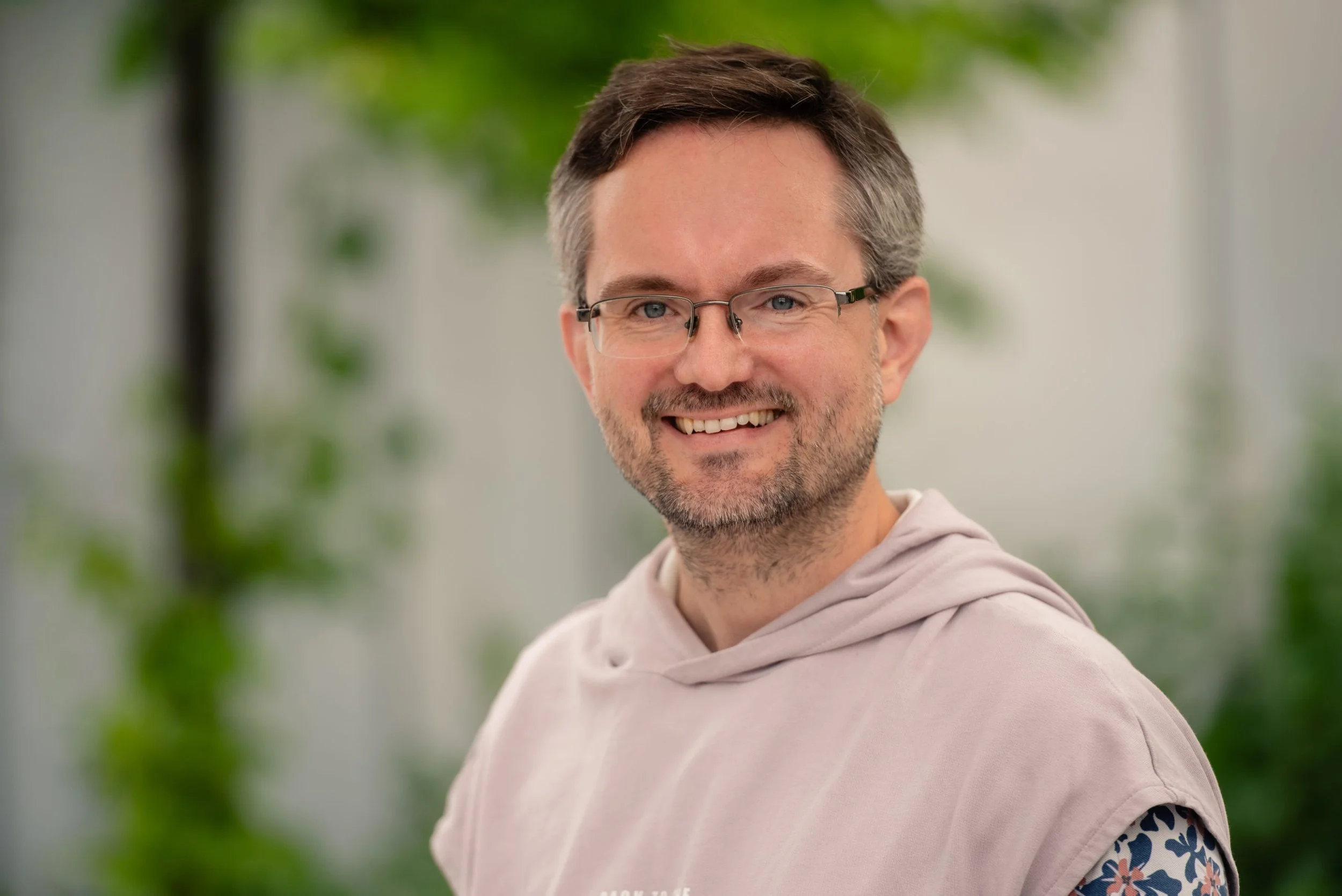 A smiling man wearing glasses and a light-colored hoodie, standing outdoors with blurred green foliage in the background.