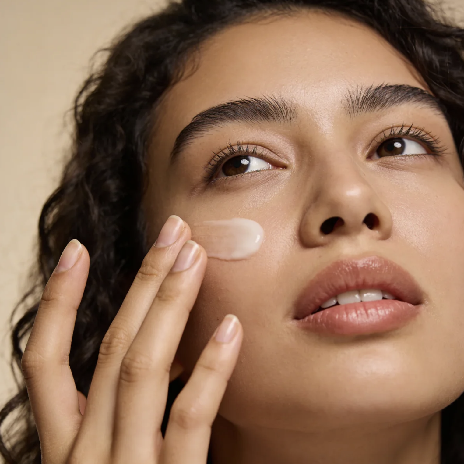 A woman with curly hair applying a skincare product to her cheek.