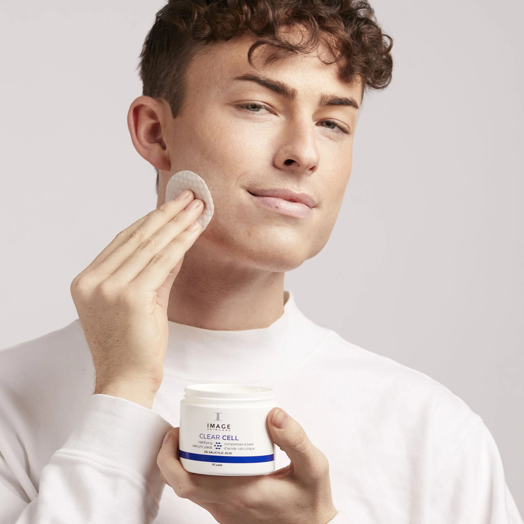 A young man with curly brown hair applying a skincare product to his cheek using a cotton pad. He is holding a white jar of skincare cream labeled 'Image Skincare Clear Cell' with both hands, and wearing a white shirt. The background is plain and light-colored.