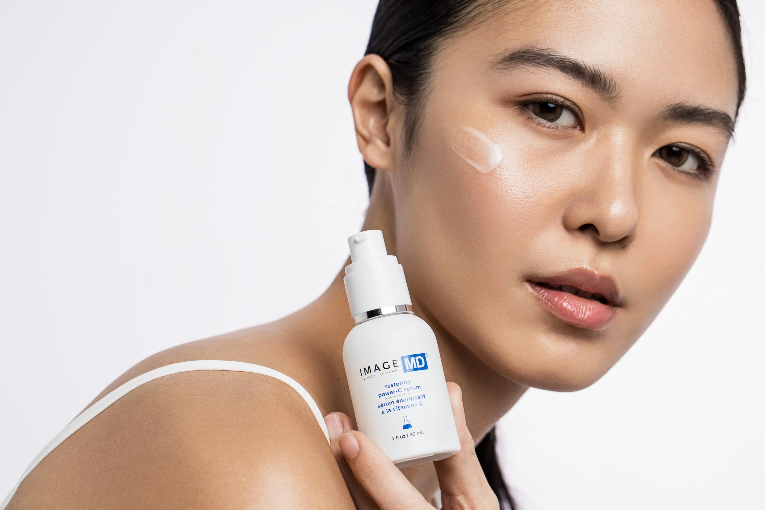 A woman with dark hair and makeup applying skincare, holding a bottle labeled 'Image MD' with a white cap and blue text, against a plain light background.