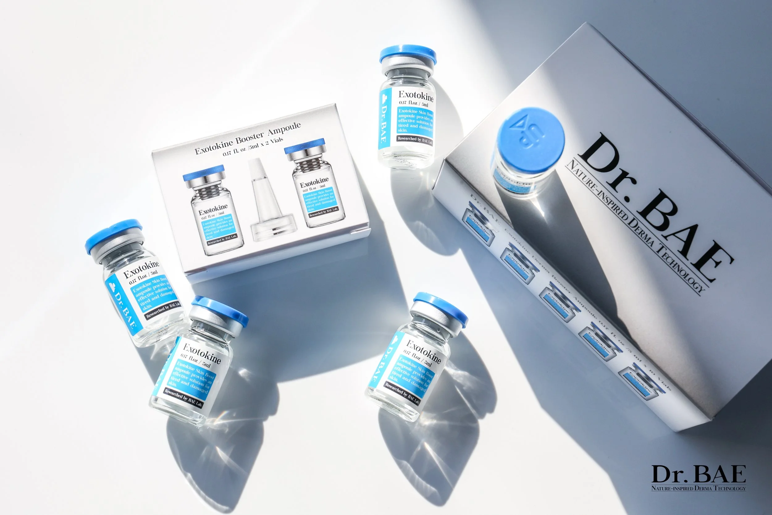 Several small bottles labeled 'Exotokine' with blue caps, scattered near a white box with similar branding and text, on a white surface with shadows.