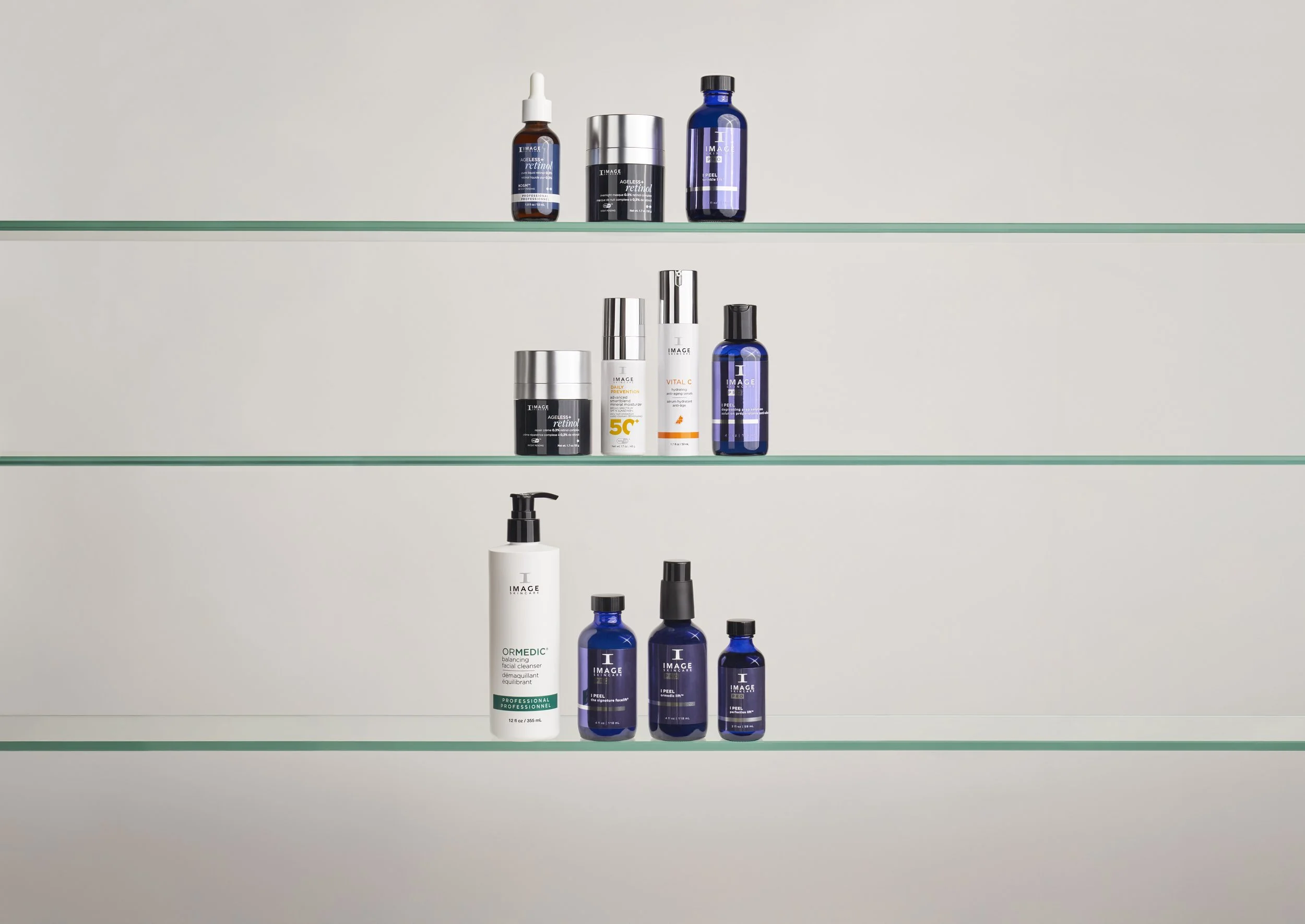 Three glass shelves with various skincare products, including bottles with pumps, dropper bottles, and jars, mostly in blue, white, and silver packaging, displayed against a plain background.