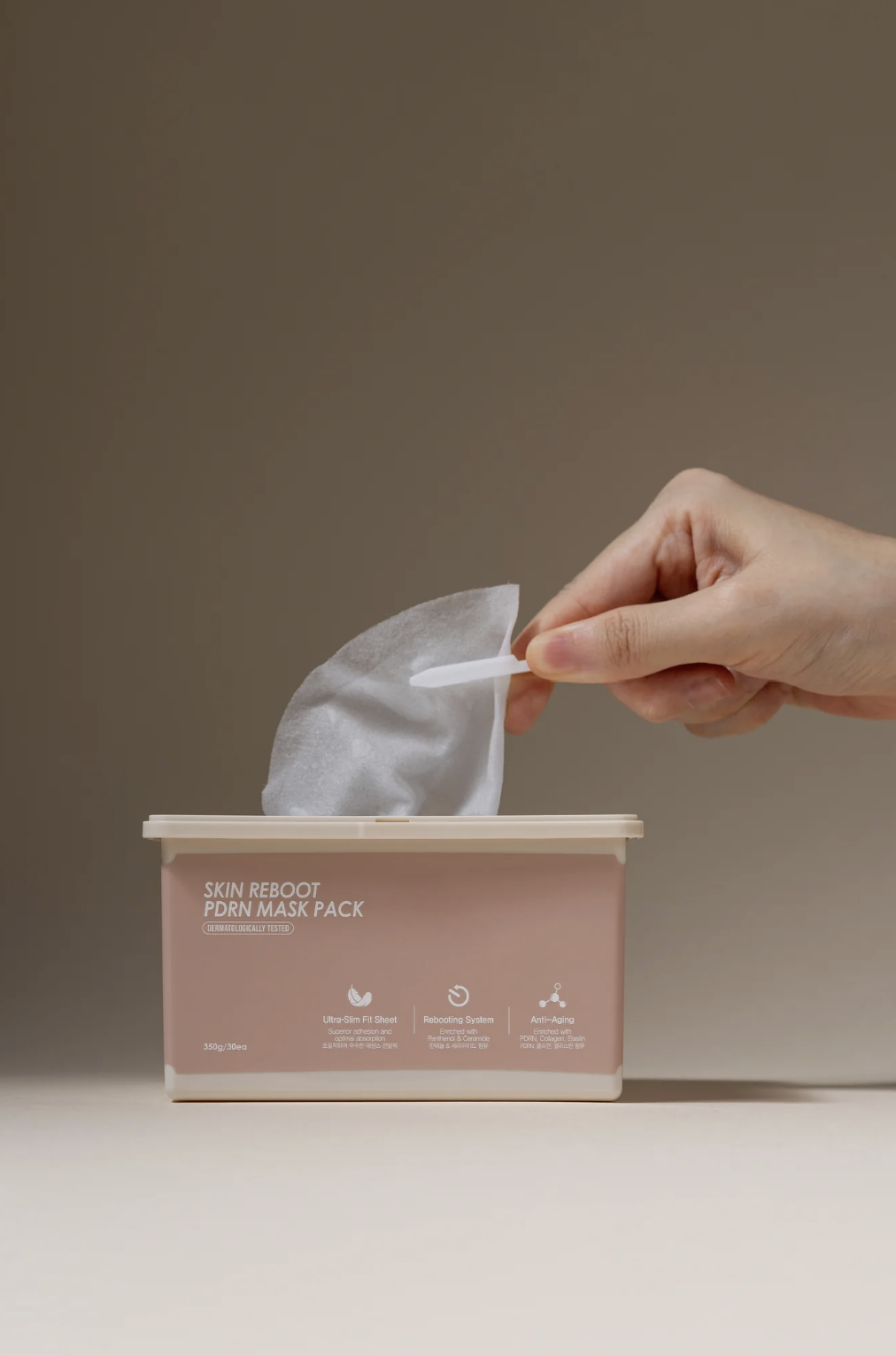 A hand removing a sheet mask from a pink box labeled 'Skin Reboot PDRN Mask Pack' with icons and text about its features.