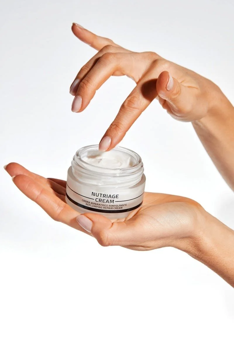 A person's hand holding a jar of Nutriage cream while another hand dips a finger into the cream.