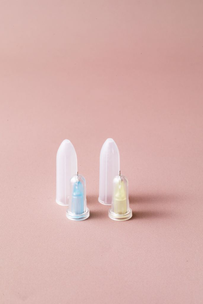 Two sets of small nasal spray bottles with clear caps, one containing blue liquid and the other yellow, arranged on a pink surface against a pink background.