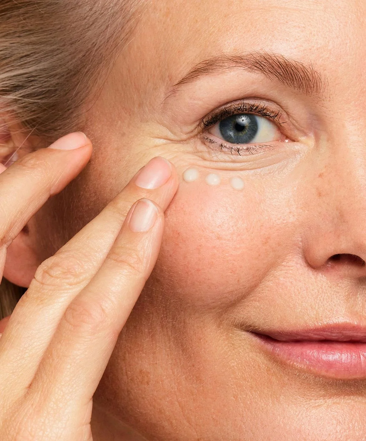 Close-up of a woman applying a skincare product under her eye using her fingers. The woman has blue eyes, fair skin, and light brown hair.