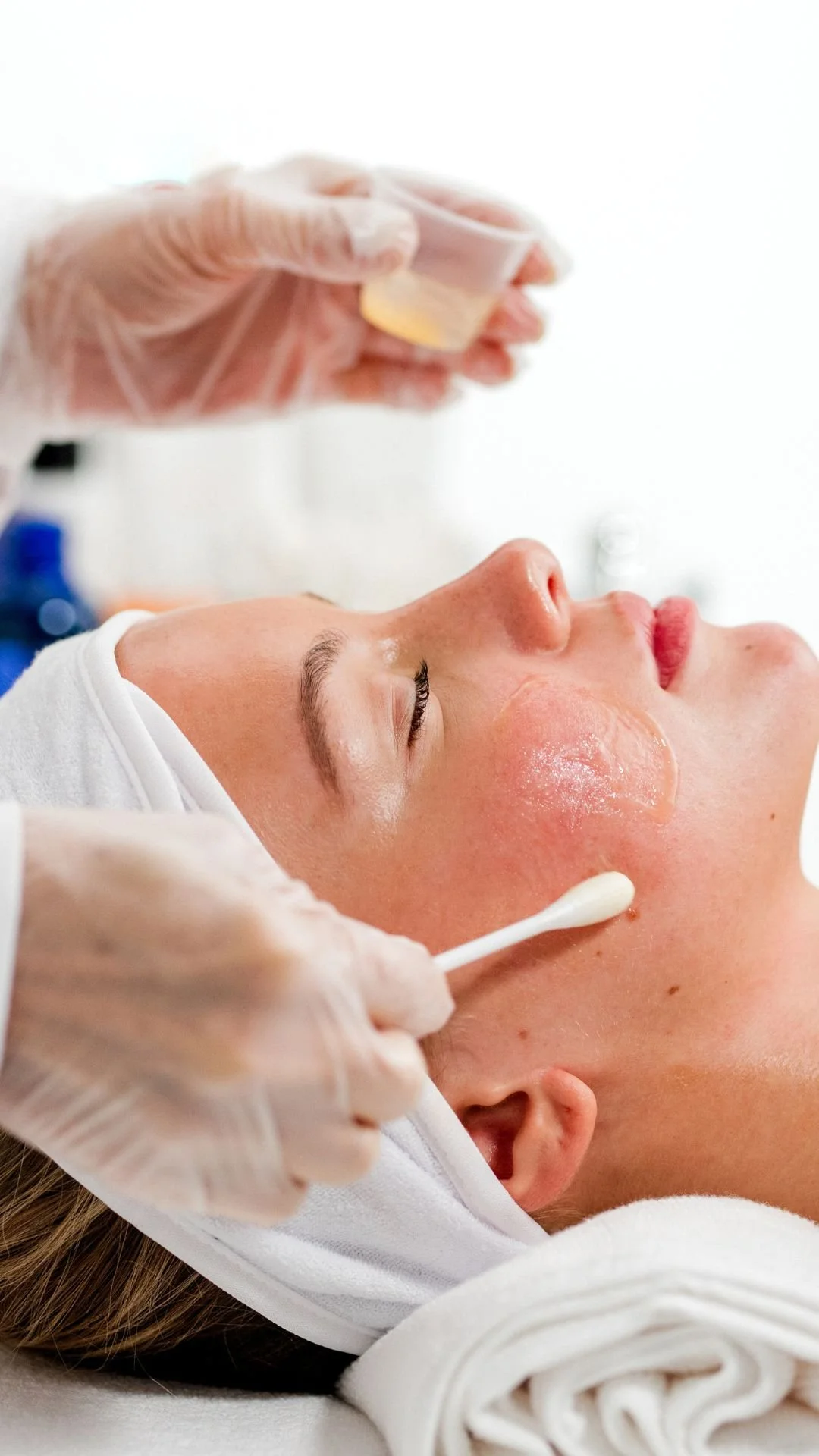 A woman receives a facial treatment at a spa, lying down with her eyes closed while a skincare specialist applies a product with a cotton swab to her face.